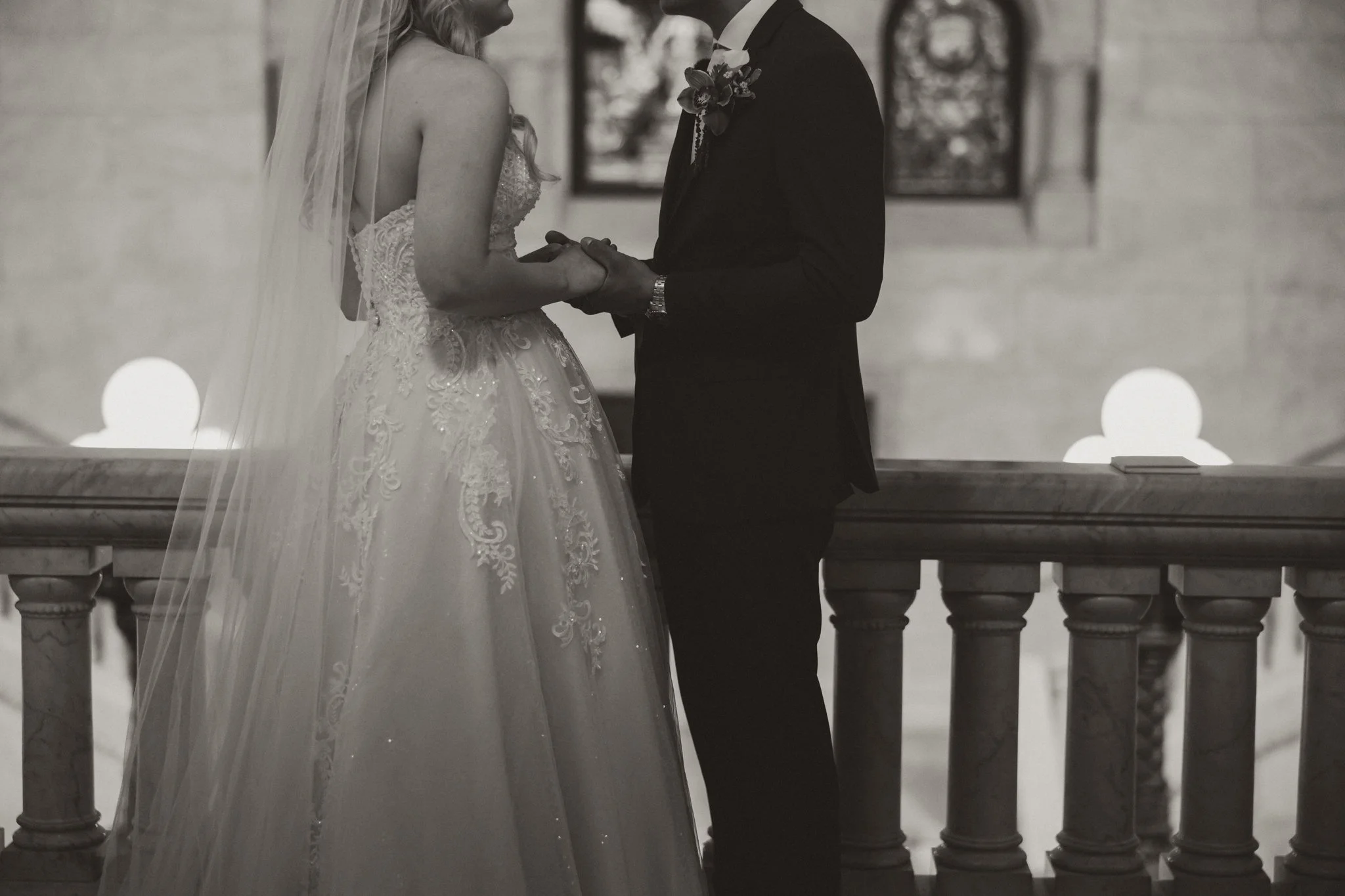 Black and white photo of a bride and groom holding hands during a wedding ceremony inside a church or chapel.