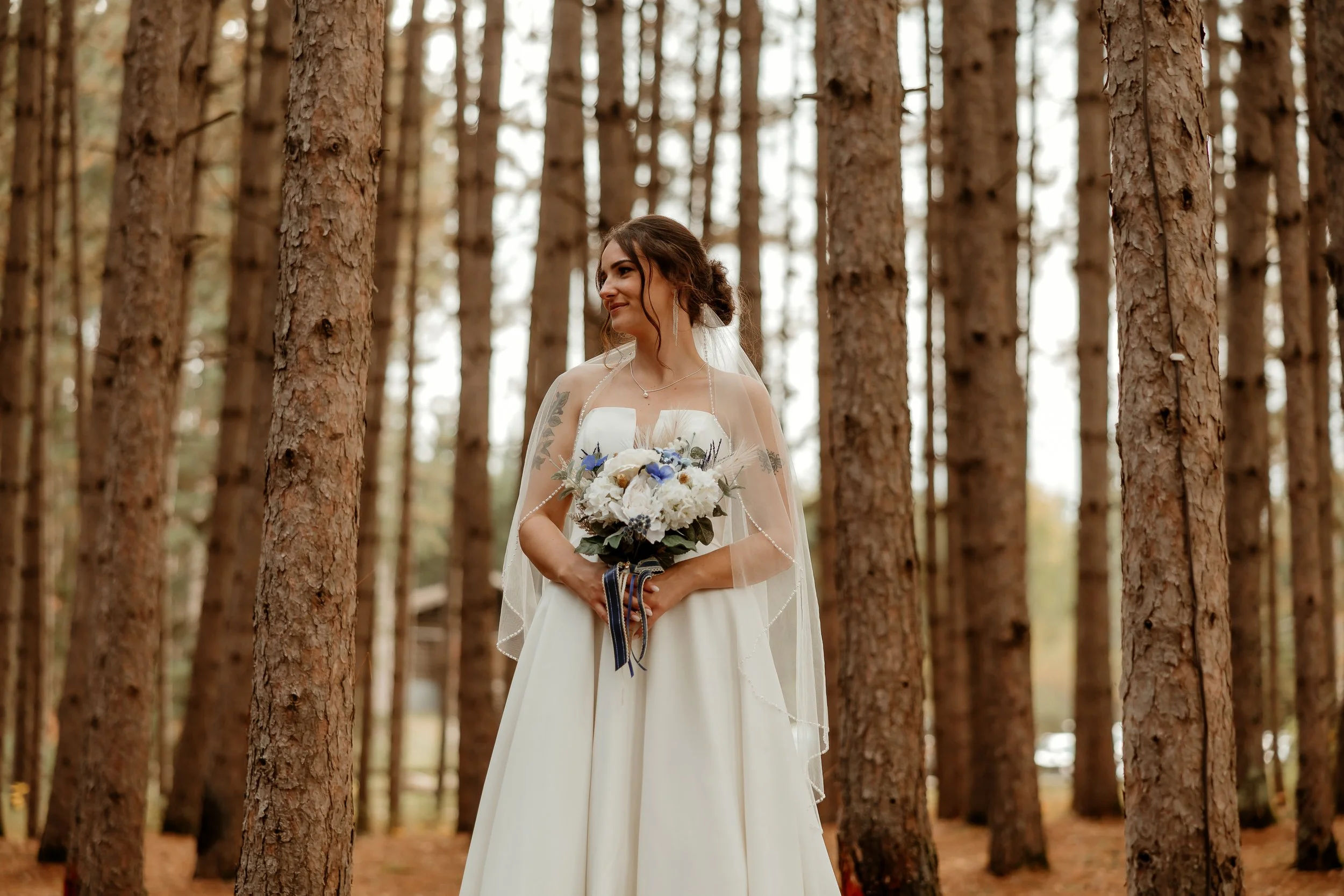 A bride holding a bouquet in a forest with tall, straight trees and soft natural lighting.