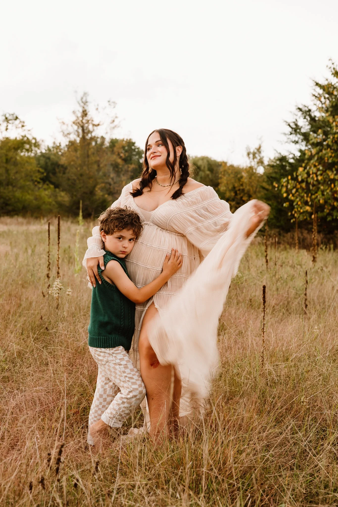 A pregnant woman in a flowing beige dress standing in a grassy field with a young boy hugging her, surrounded by trees, during daytime.