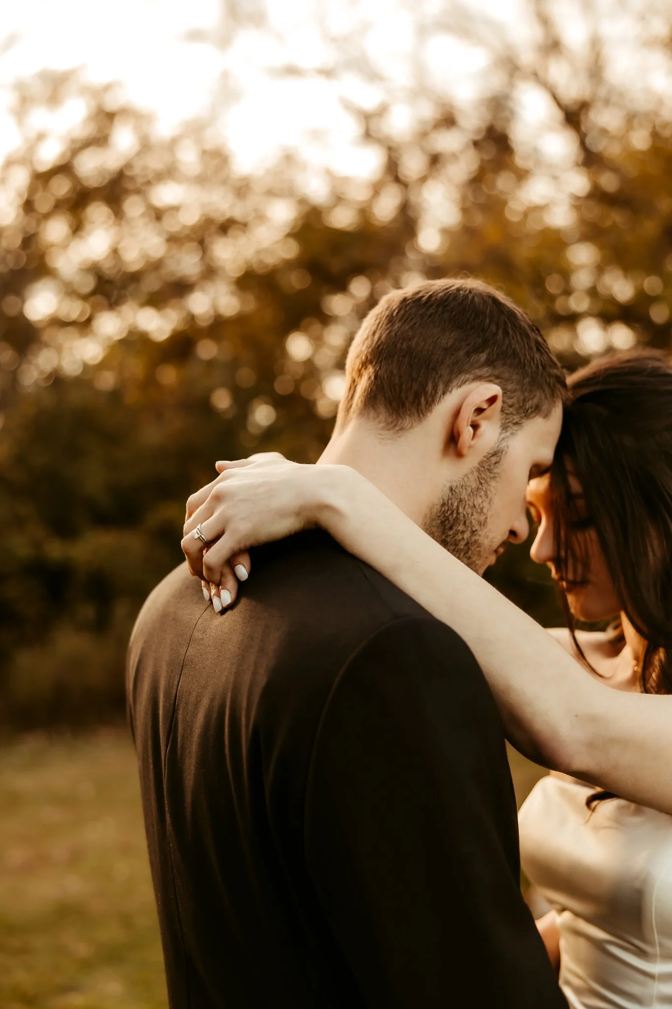 A couple touching foreheads outdoors during sunset, with trees and warm lighting in the background.