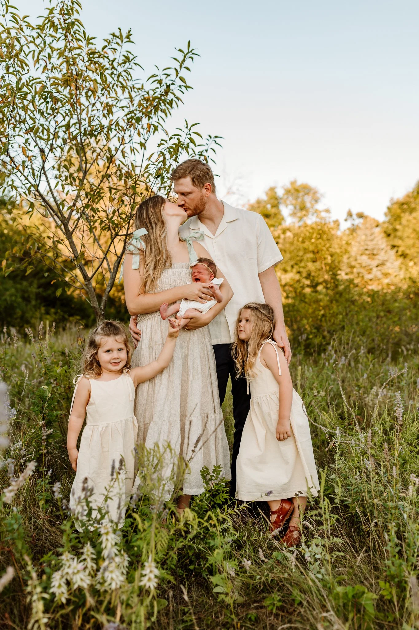 A family of six standing together outdoors in a field during sunset, with the parents kissing and the children around them, including a newborn being held by the mother.
