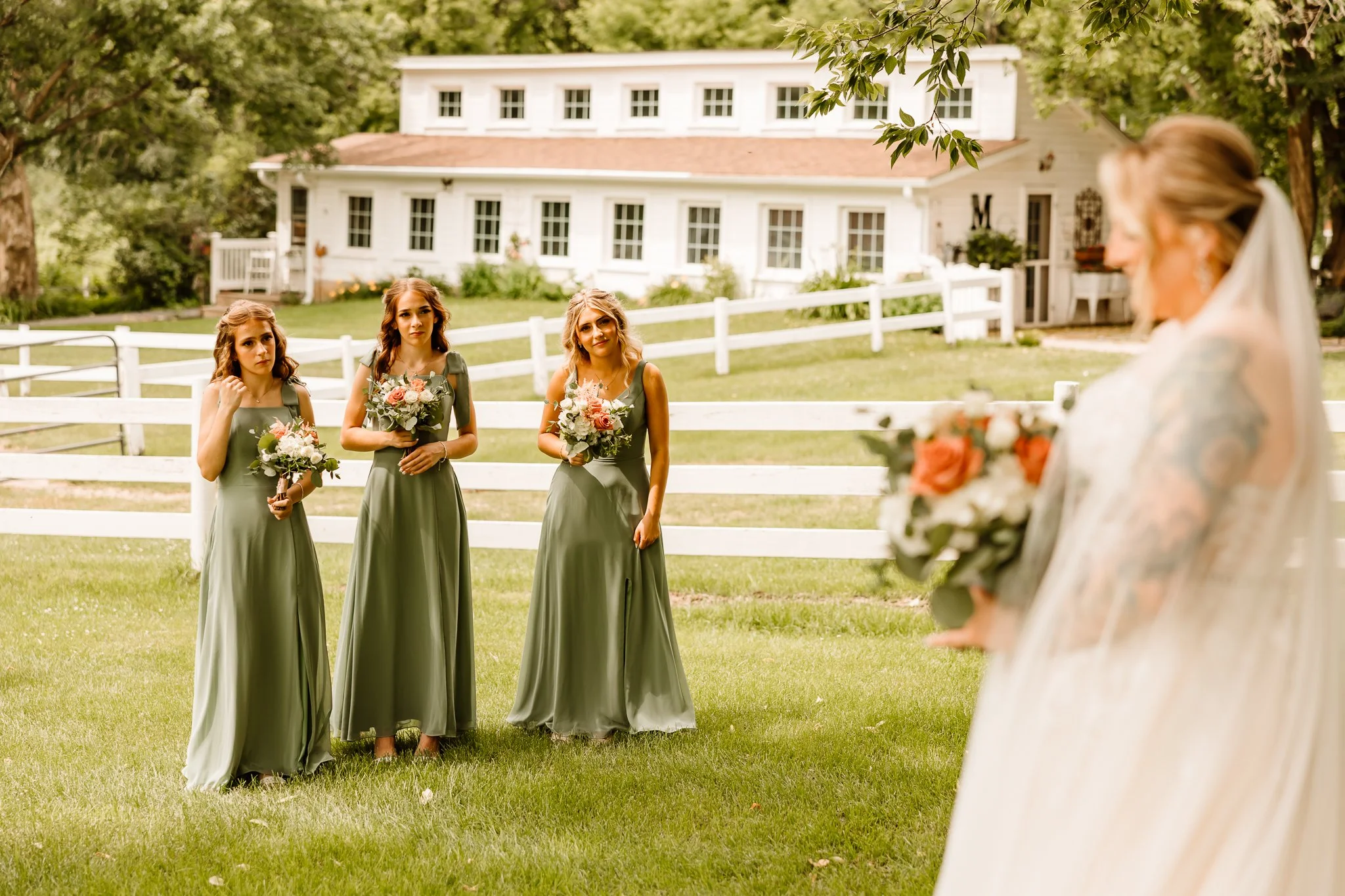A bride holding a bouquet looks at three bridesmaids, also holding bouquets, standing in a grassy area outdoors near a white fence and a large white house.
