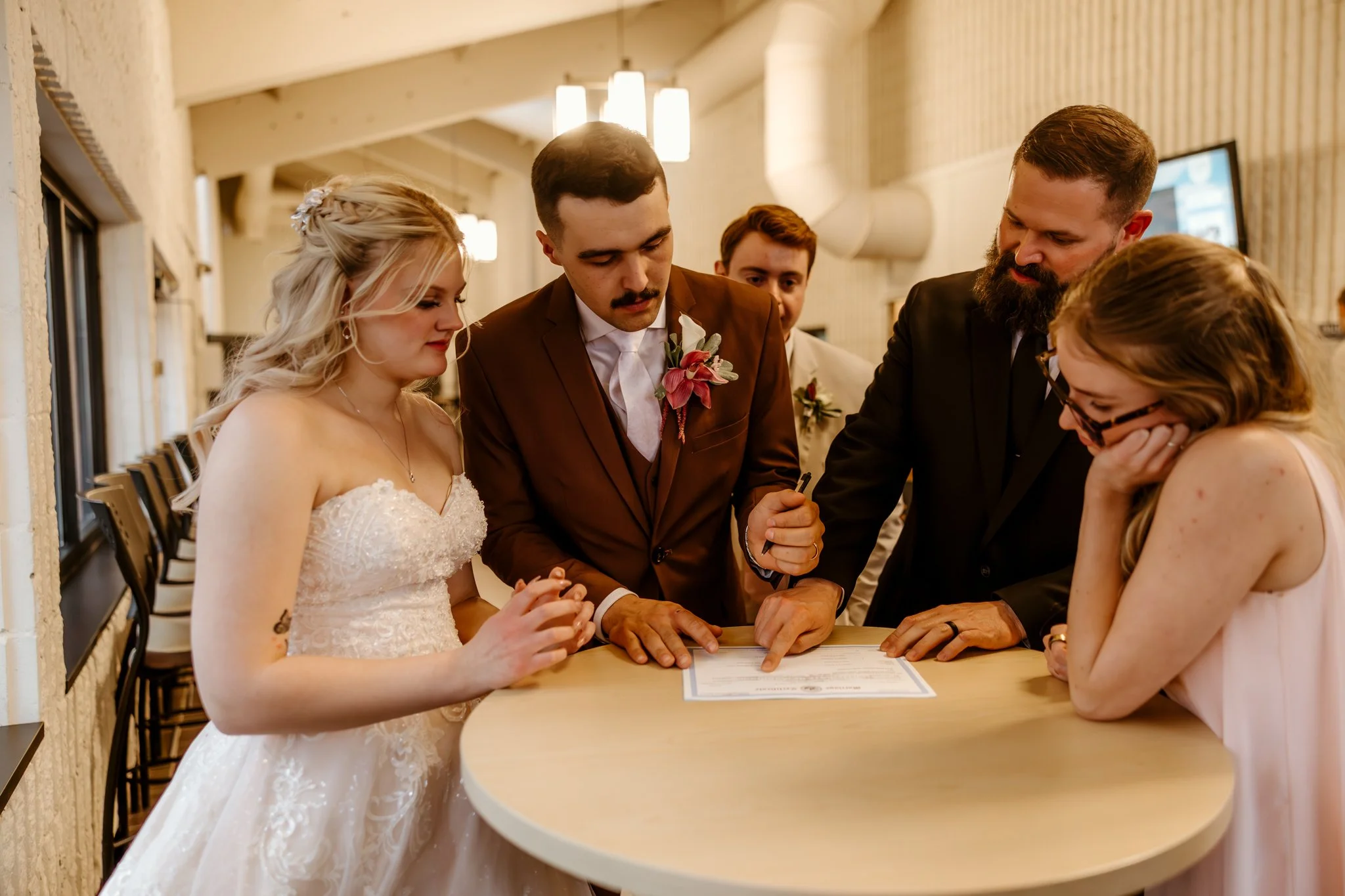 People gathered around a table, signing documents, dressed in wedding attire during a wedding ceremony.
