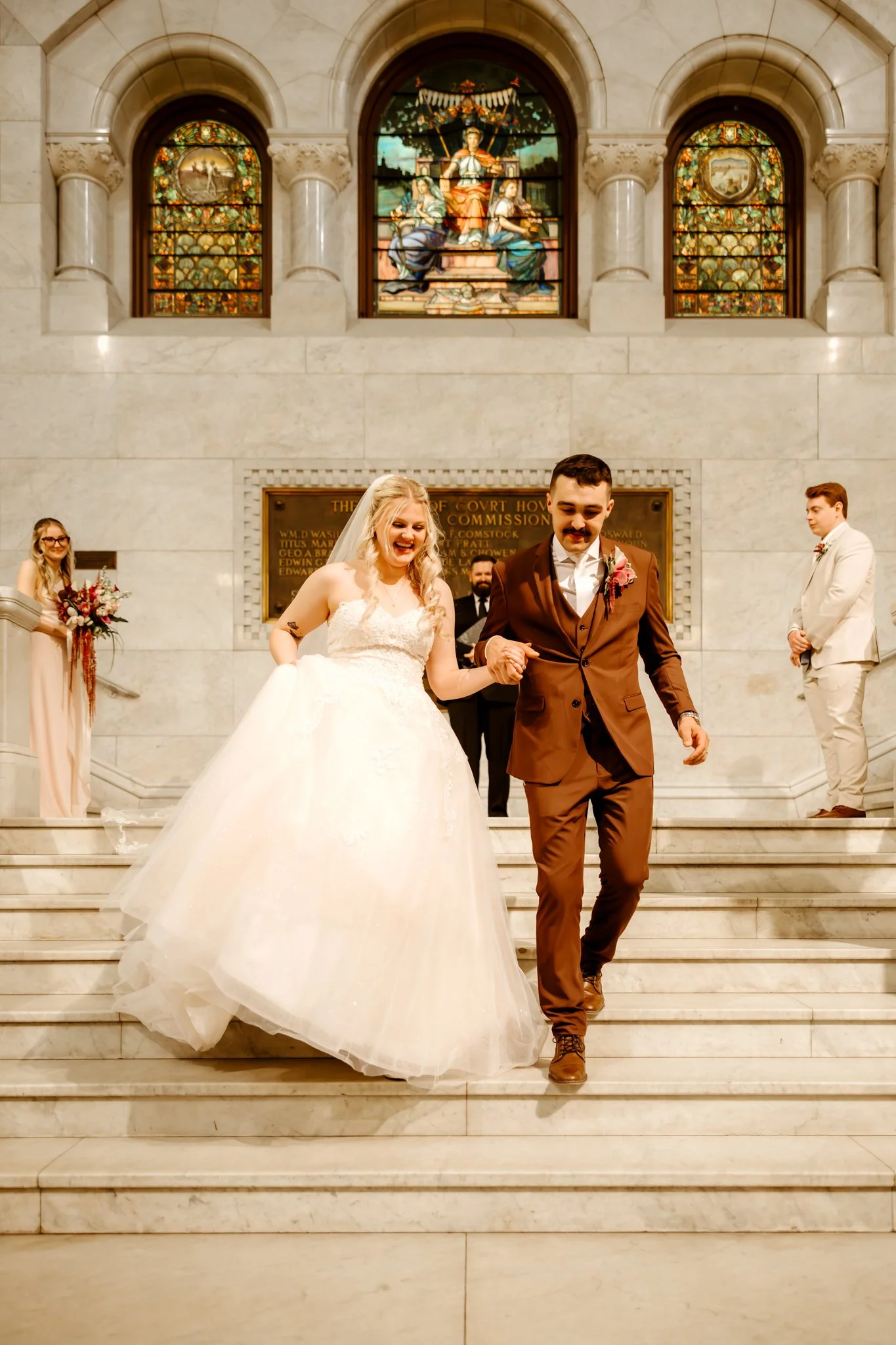 A newly married couple descending the stairs inside a courthouse or cathedral, with the bride in a white wedding dress and the groom in a brown suit, surrounded by their wedding party and guests.