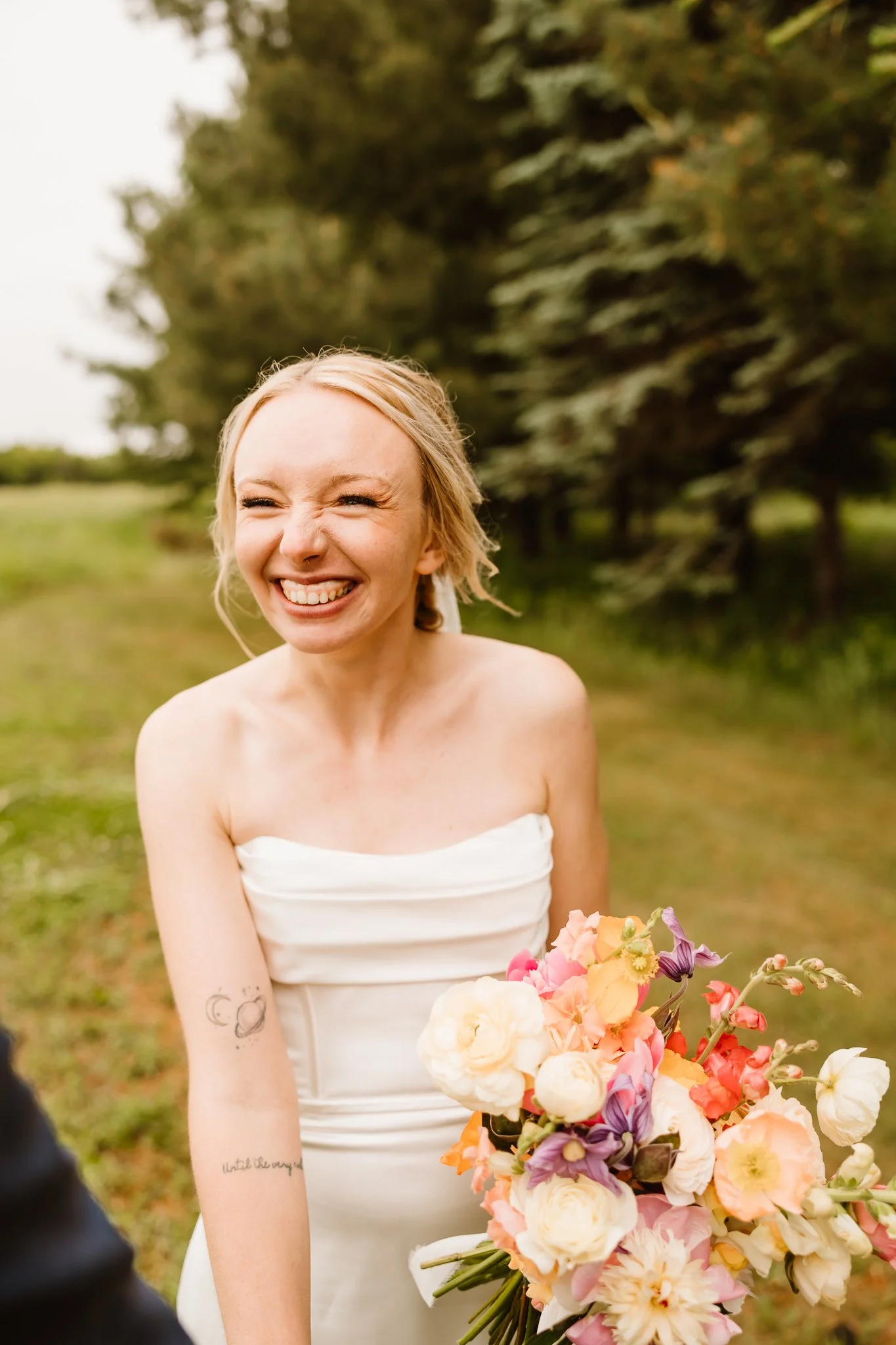 A smiling woman in a white strapless dress holding a bouquet of colorful flowers outdoors.