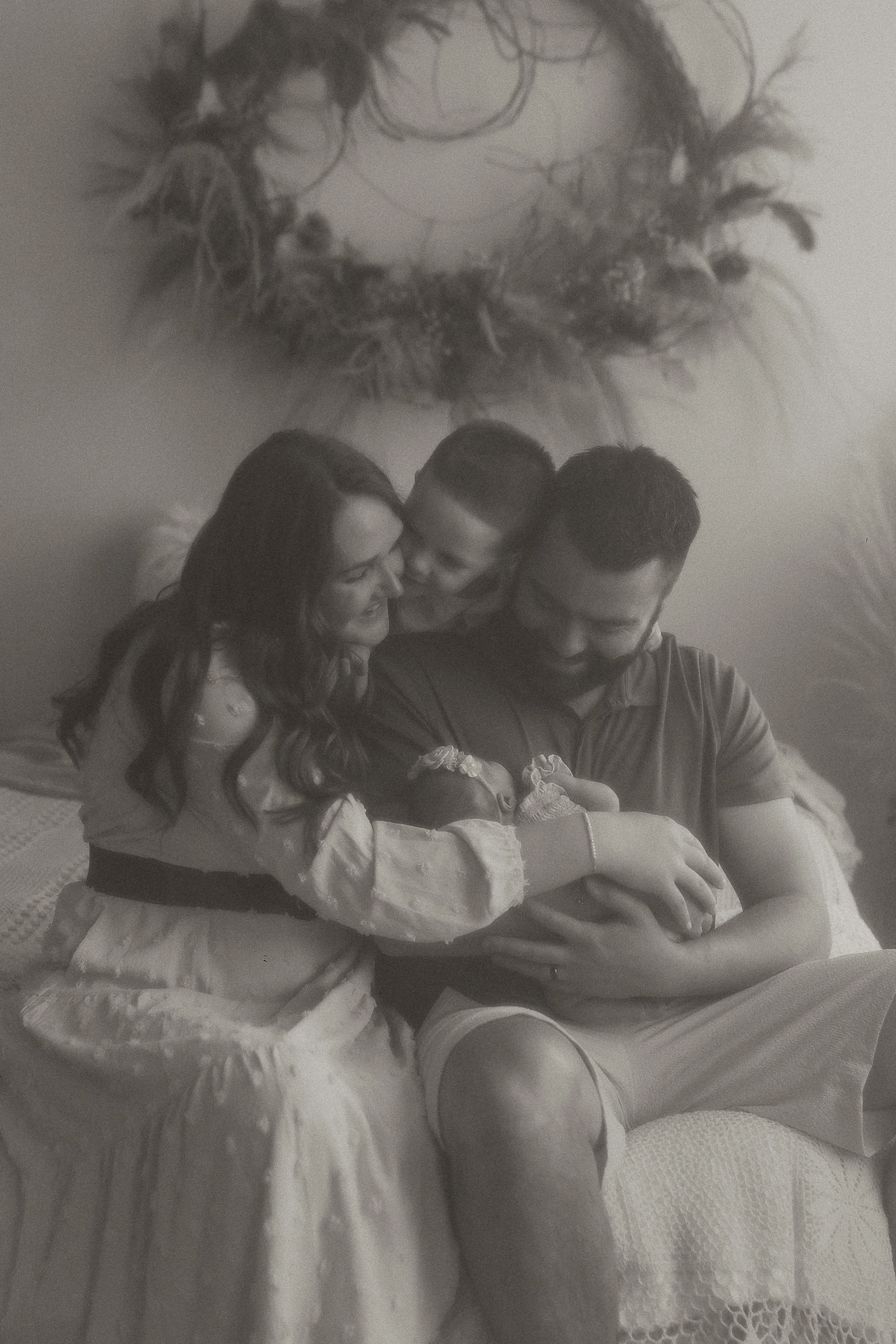A family of four sitting on a couch, with two newborn twins in their arms, smiling and looking at the babies. A wreath made of twigs is hanging on the wall behind them.