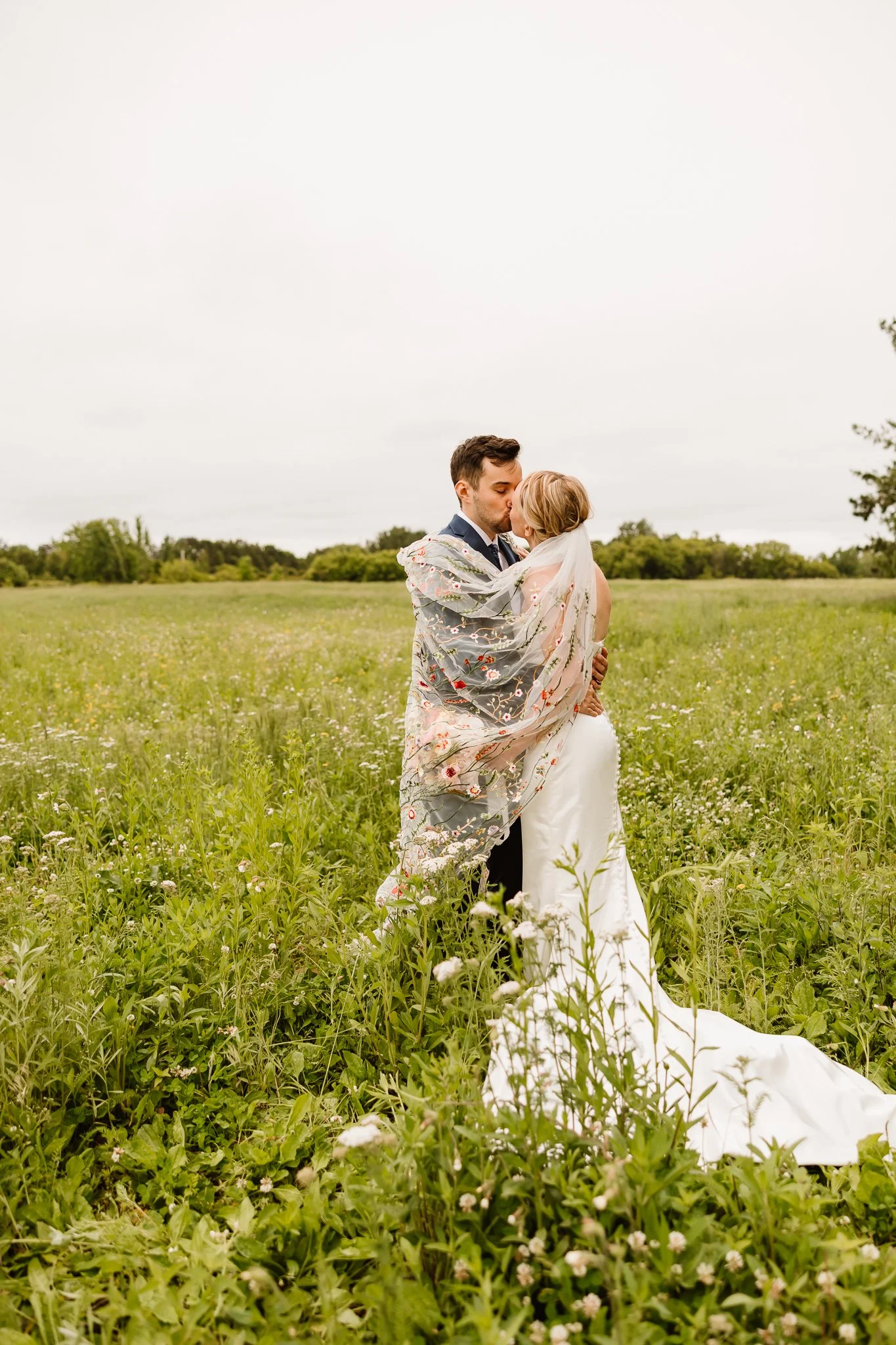 A newlywed couple sharing a kiss in a grassy field, with the groom wrapped in a floral shawl and the bride wearing a white wedding dress and veil, under an overcast sky.