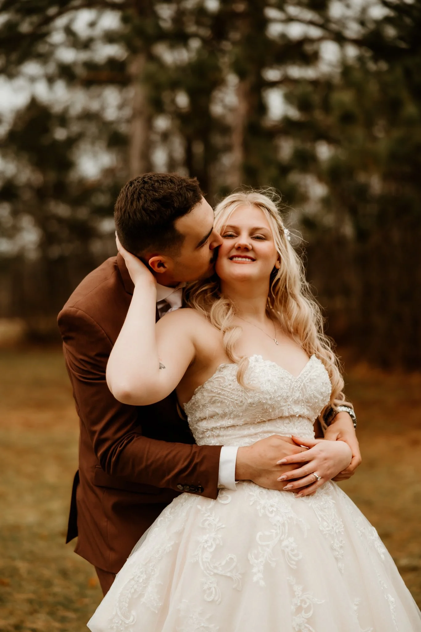 A newlywed couple outdoors, with the man kissing the woman's cheek as she smiles, wearing a white wedding gown and brown suit, with trees in the background.