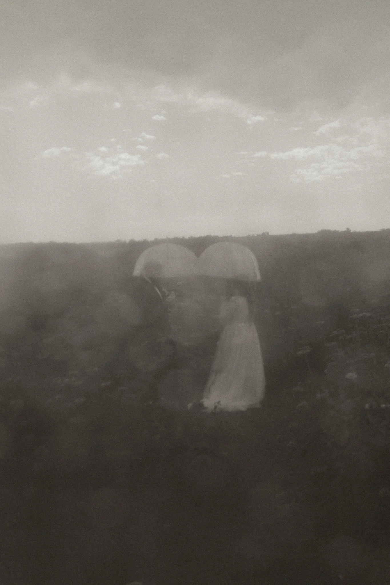 A black-and-white photo of two people, a bride and groom, holding umbrellas and walking on a rocky landscape with a cloudy sky in the background.