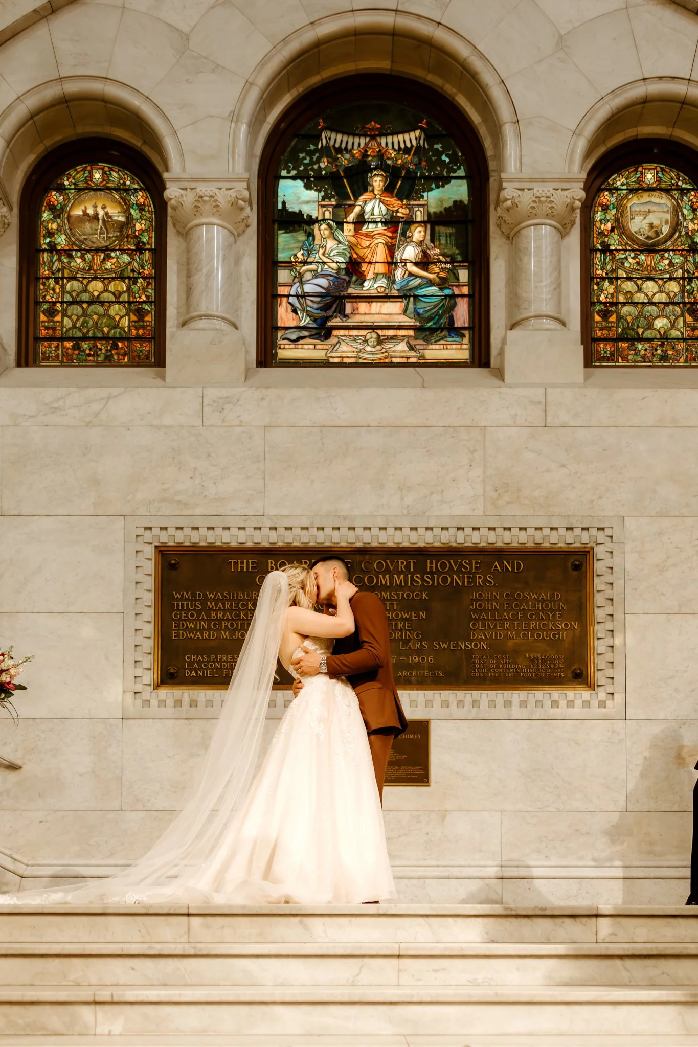 A bride and groom sharing a kiss during their wedding inside a church with stained glass windows and a plaque on the wall.