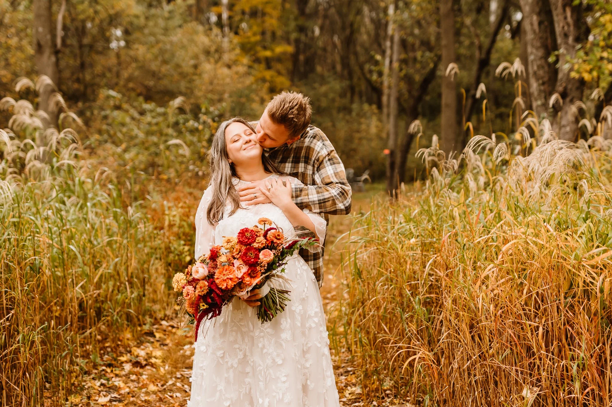A couple embraces in a field of tall plants with fall foliage in the background. The woman holds a large bouquet of sunflowers, zinnias, and other flowers, and wears a white lace dress. The man leans in to kiss her on the cheek.