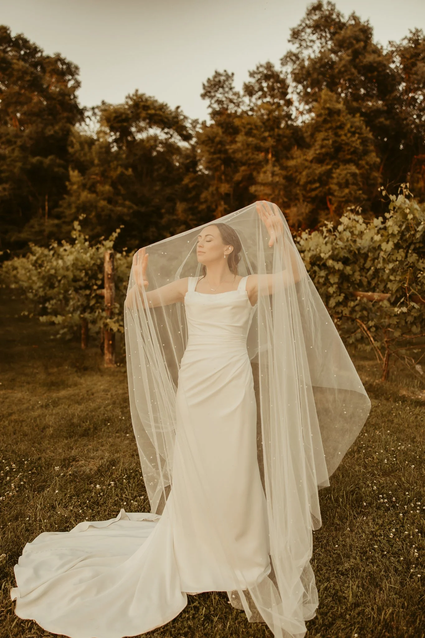 Bride standing outdoors wearing a white wedding dress, with long veil, her eyes closed and arms raised, surrounded by trees at sunset.