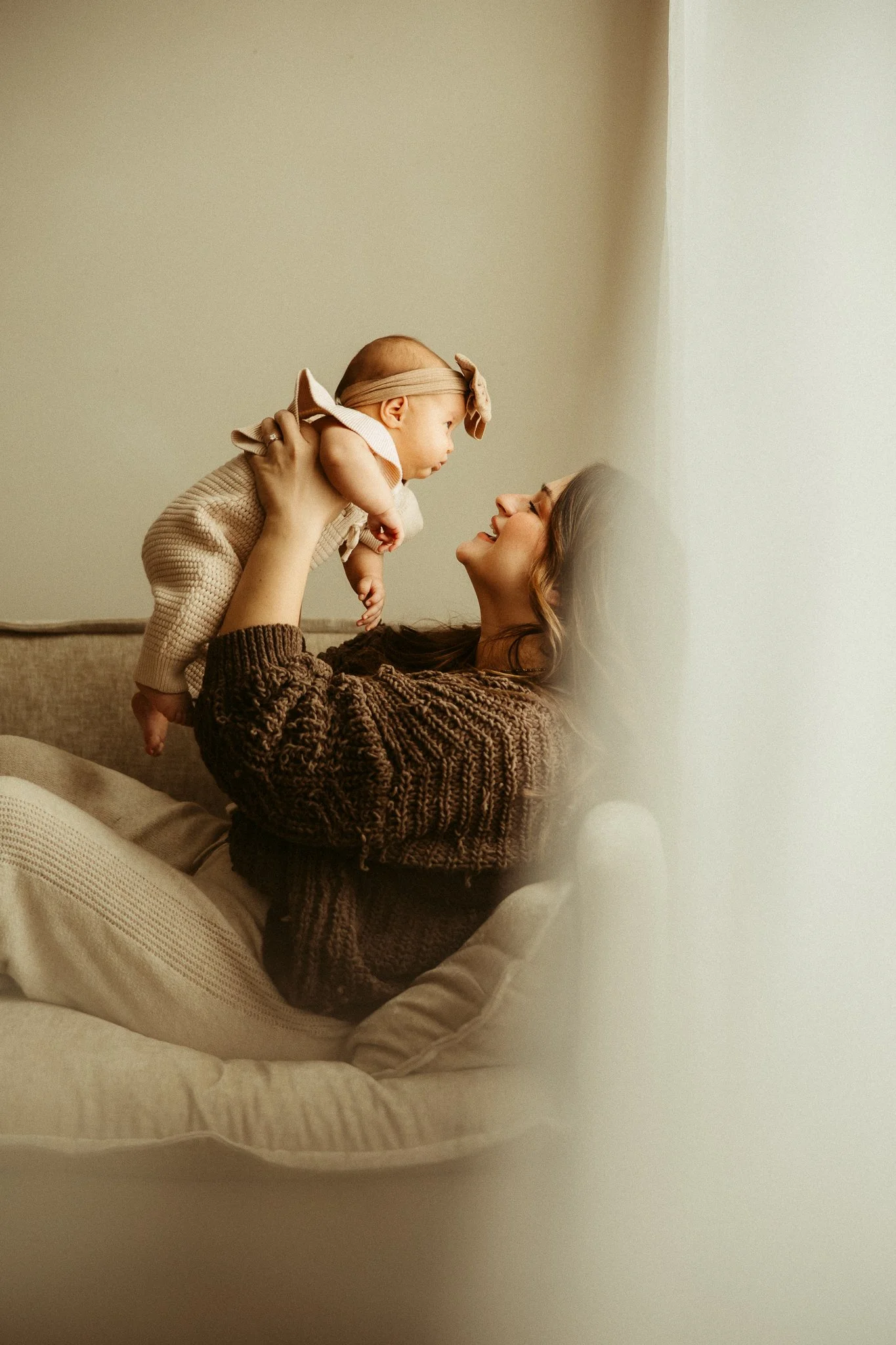 A woman holding a baby in the air, both smiling warmly, in a cozy room with soft lighting.