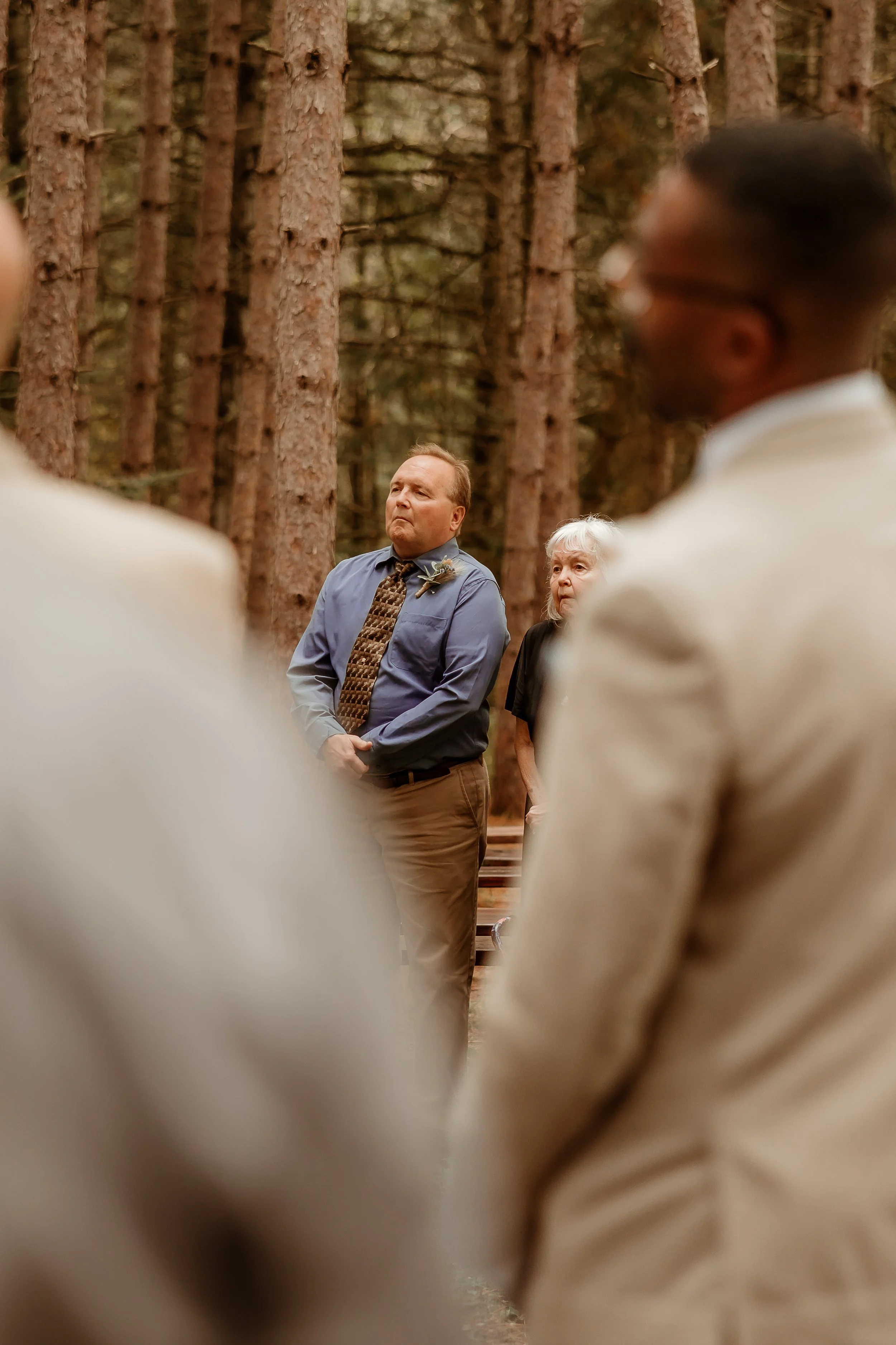People standing outdoors in a forest during a wedding ceremony, with some individuals in focus and others blurred in the foreground. Bigfork mn