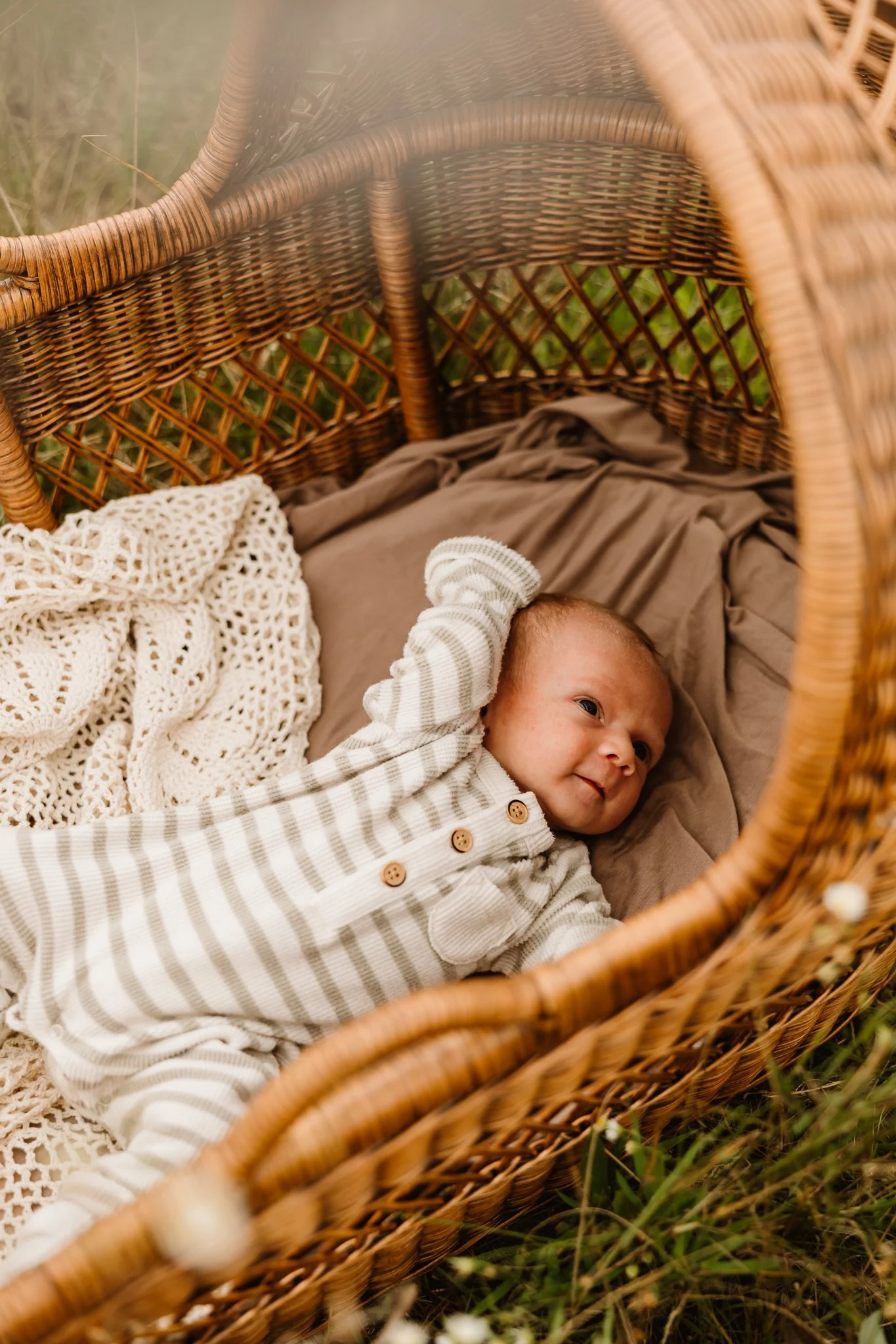 A baby lying in a wicker bassinet outdoors, covered with a beige blanket, wearing a gray and white striped onesie with buttons, and looking towards the side.