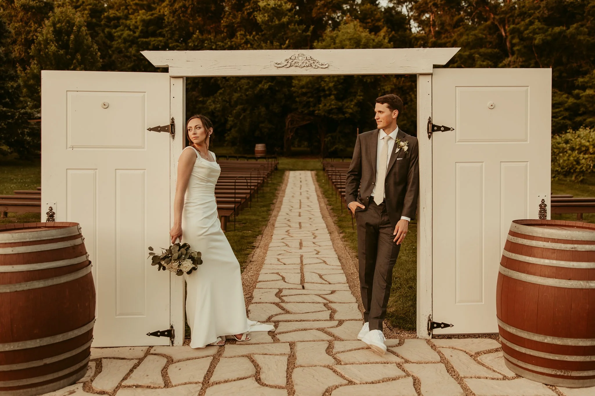 A bride and groom standing at an outdoor wedding venue, separated by a wide white gate, with trees in the background and a stone pathway between decorated barrels.