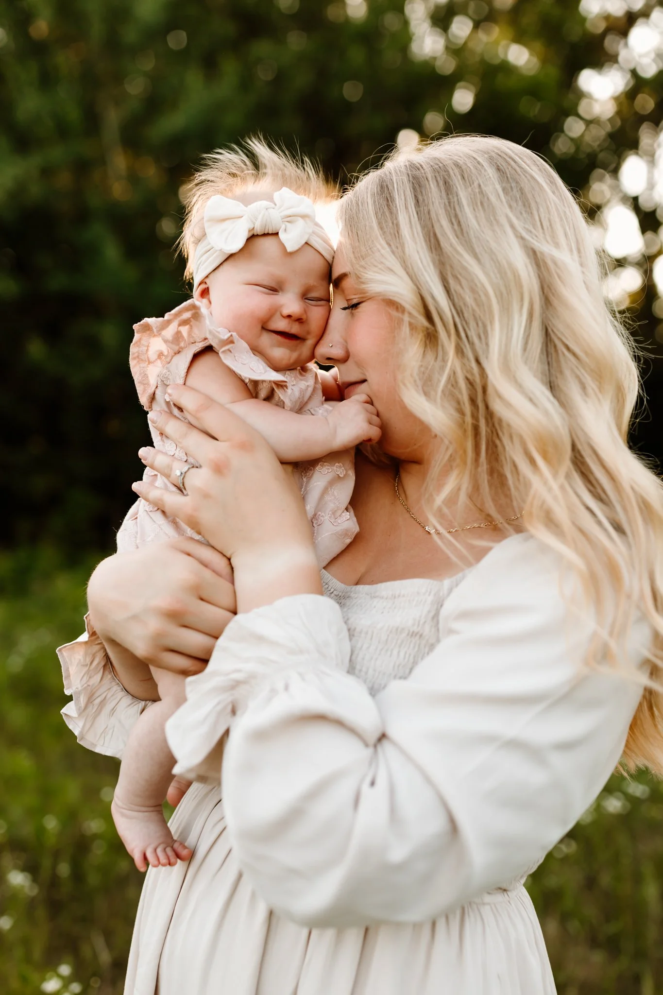 A woman holding a smiling baby girl outdoors with trees in the background, warm sunlight illuminating them.