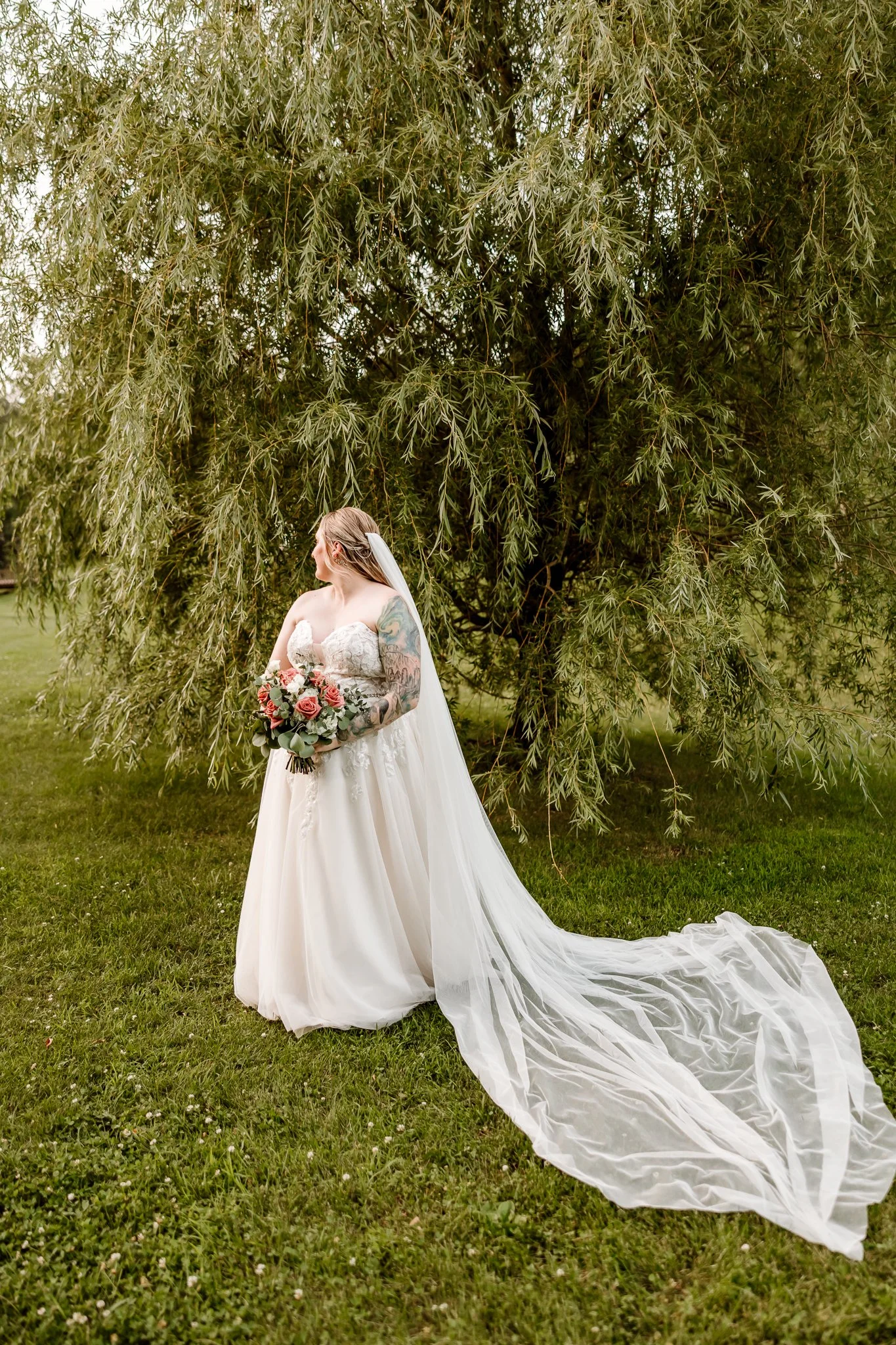 A bride in a white wedding dress holding a bouquet of pink and white roses, standing outdoors on a grassy area in front of a large leafy green tree.