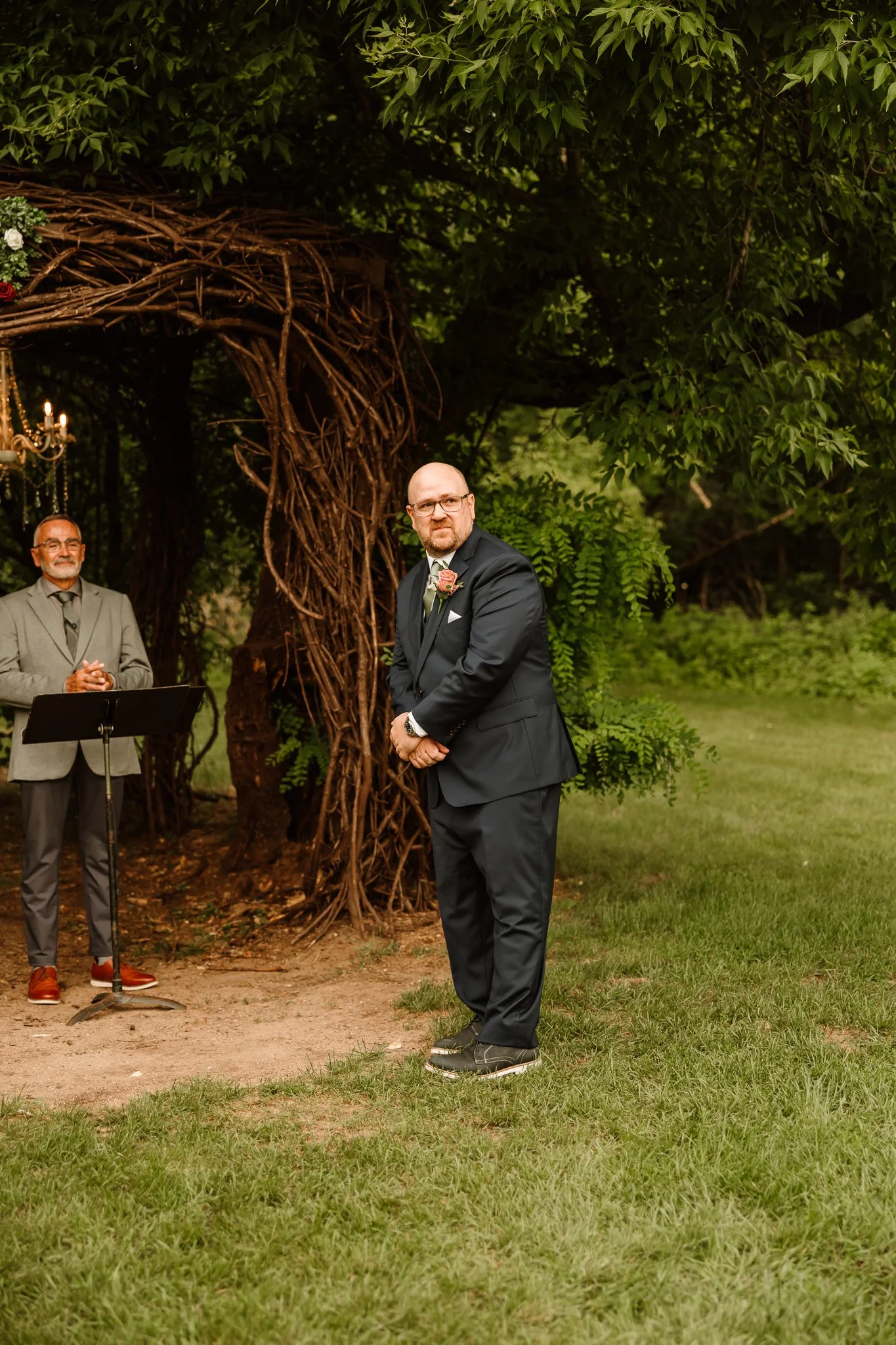 A groom stands in a garden during his wedding ceremony, facing an officiant, under a canopy of green trees with a rustic vine arch.