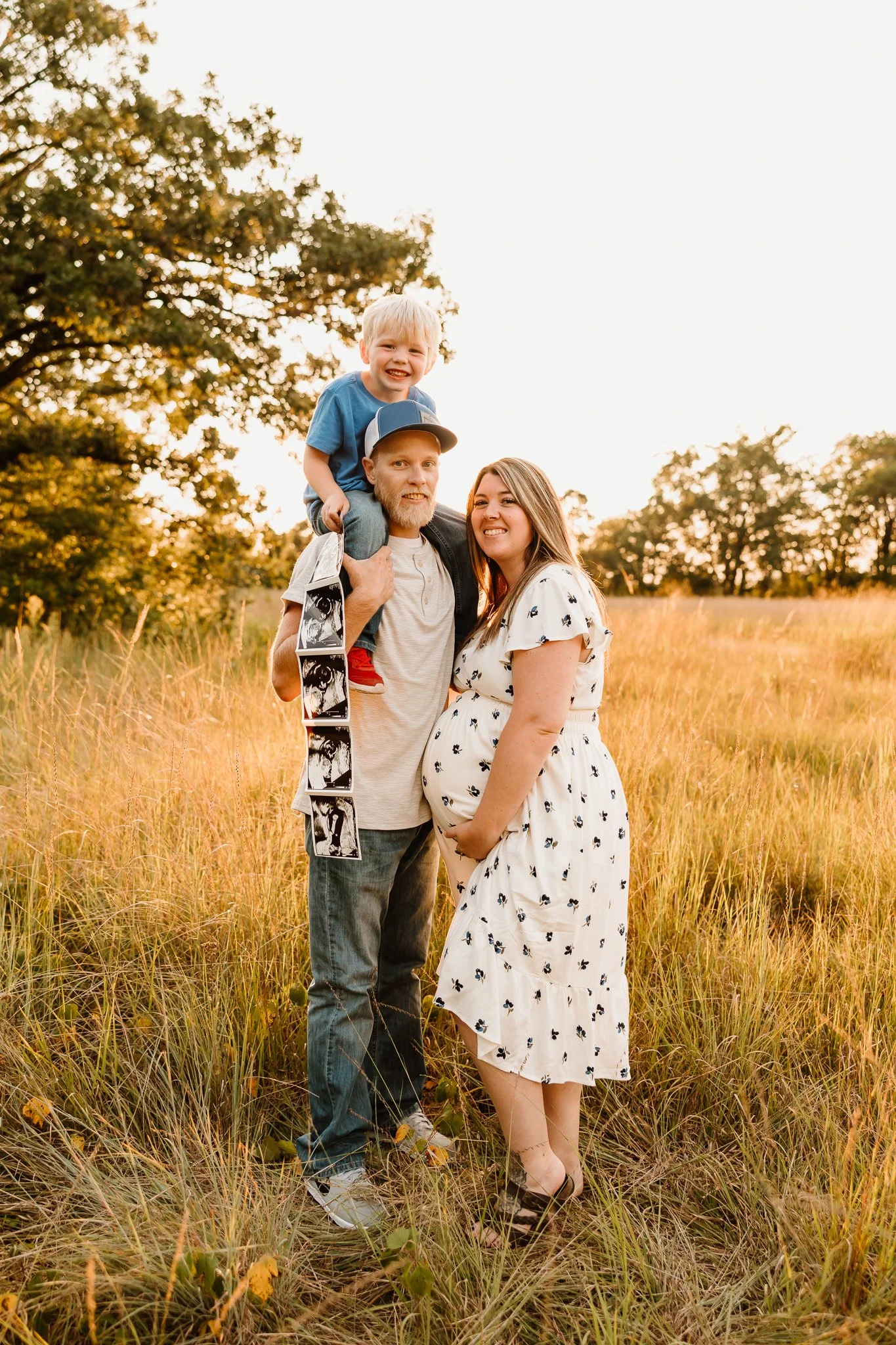 A pregnant woman, a man, and a young boy in a field at sunset. The boy is sitting on the man's shoulders, smiling and holding a strip of ultrasound images. The woman is smiling, touching her pregnant belly, and wearing a white dress with a black flor