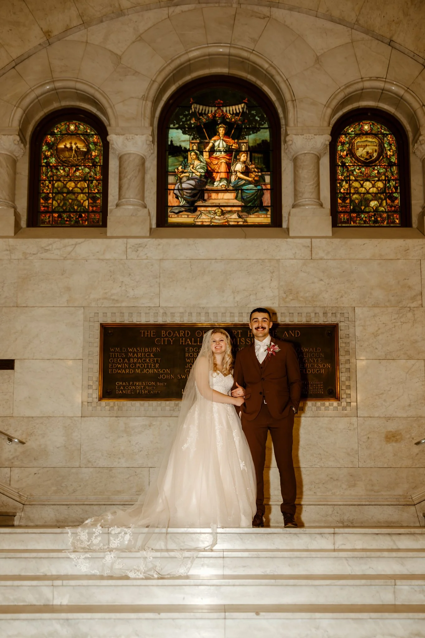 A newlywed couple dressed in wedding attire, standing on marble stairs inside a historic building with stained glass windows and a plaque on the wall behind them.