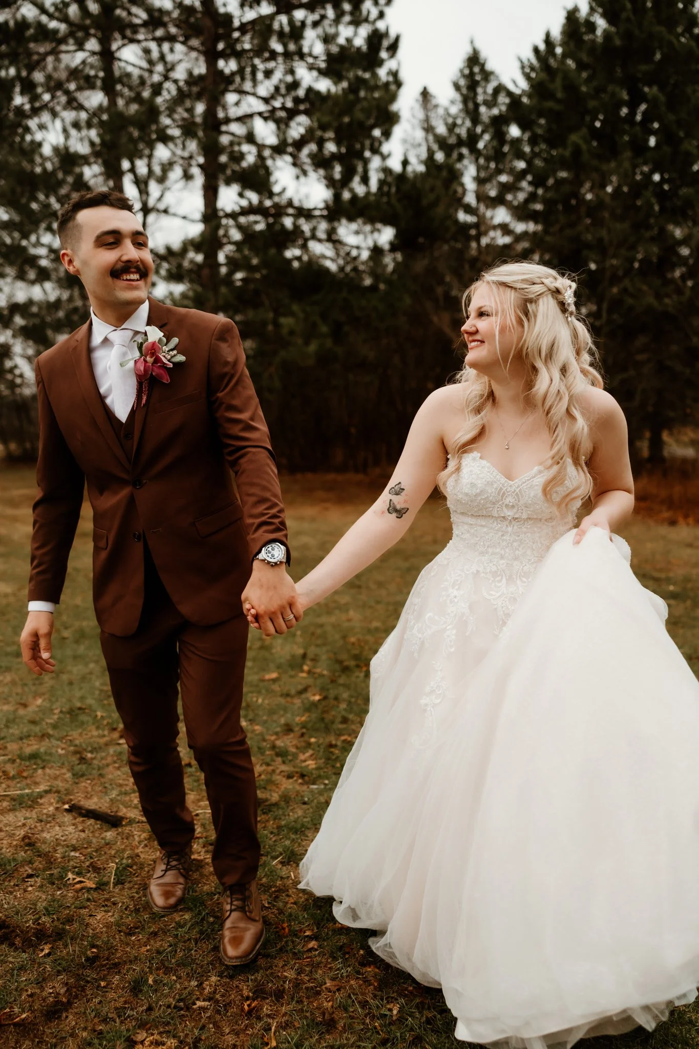 A newlywed couple holding hands and smiling outdoors on a fall day, with trees in the background. The groom is wearing a brown suit with a white shirt and a floral boutonniere, and the bride is in a white wedding dress with lace details and tattoos o