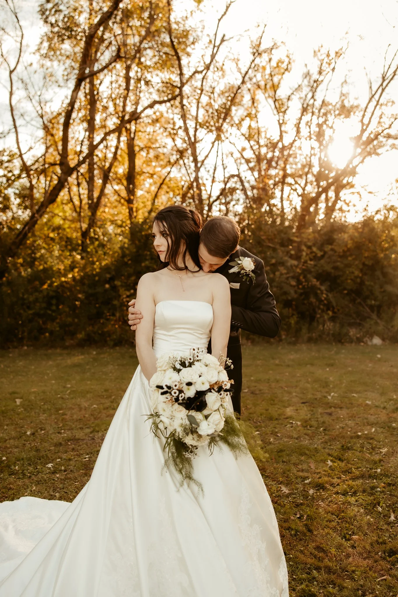 A bride and groom in wedding attire sharing a tender moment outdoors at sunset, with trees in the background and the bride holding a bouquet of white and dark flowers.