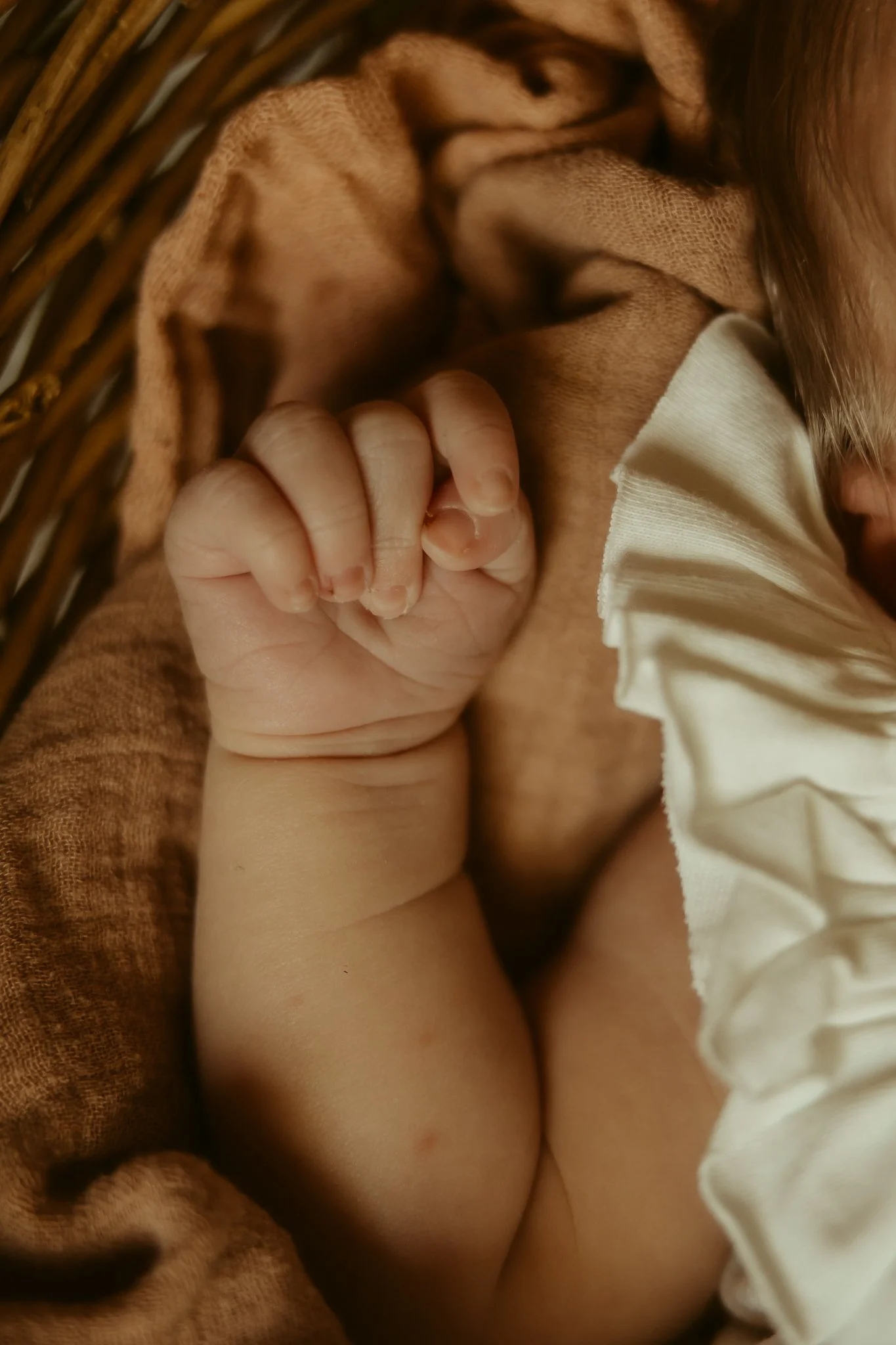 Close-up of a newborn baby lying in a basket, with a small hand clenched into a fist resting near its face, dressed in a cream-colored ruffle outfit.