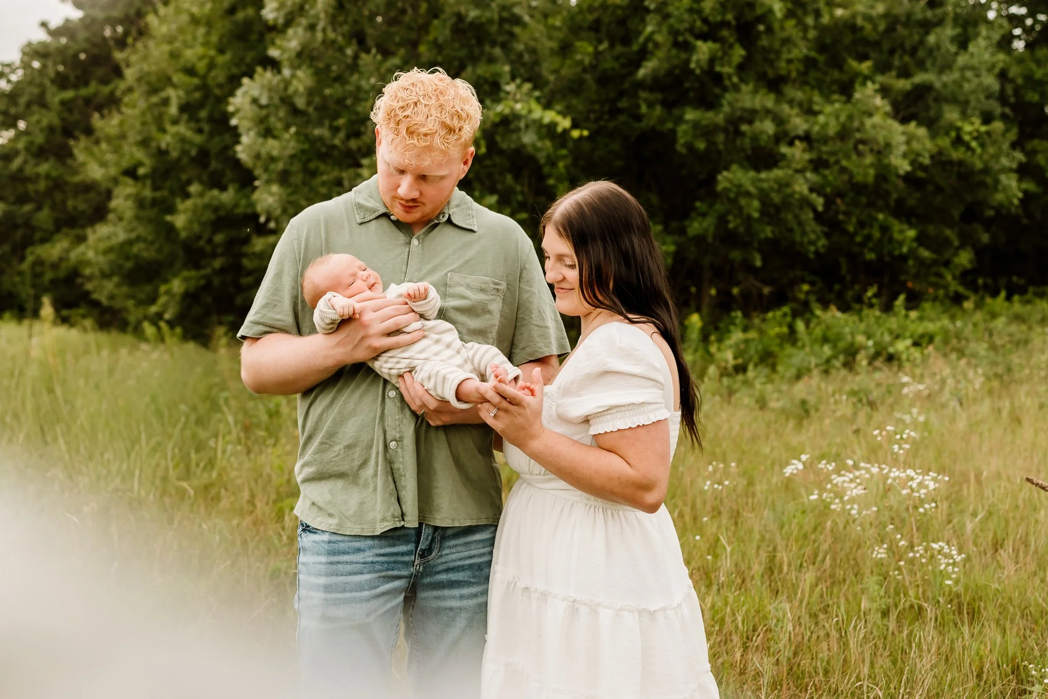 A young family of three in a grassy field with trees in the background. The father, with curly blond hair, holds a newborn baby, and the mother with long dark hair gently touches the baby while looking at it lovingly.