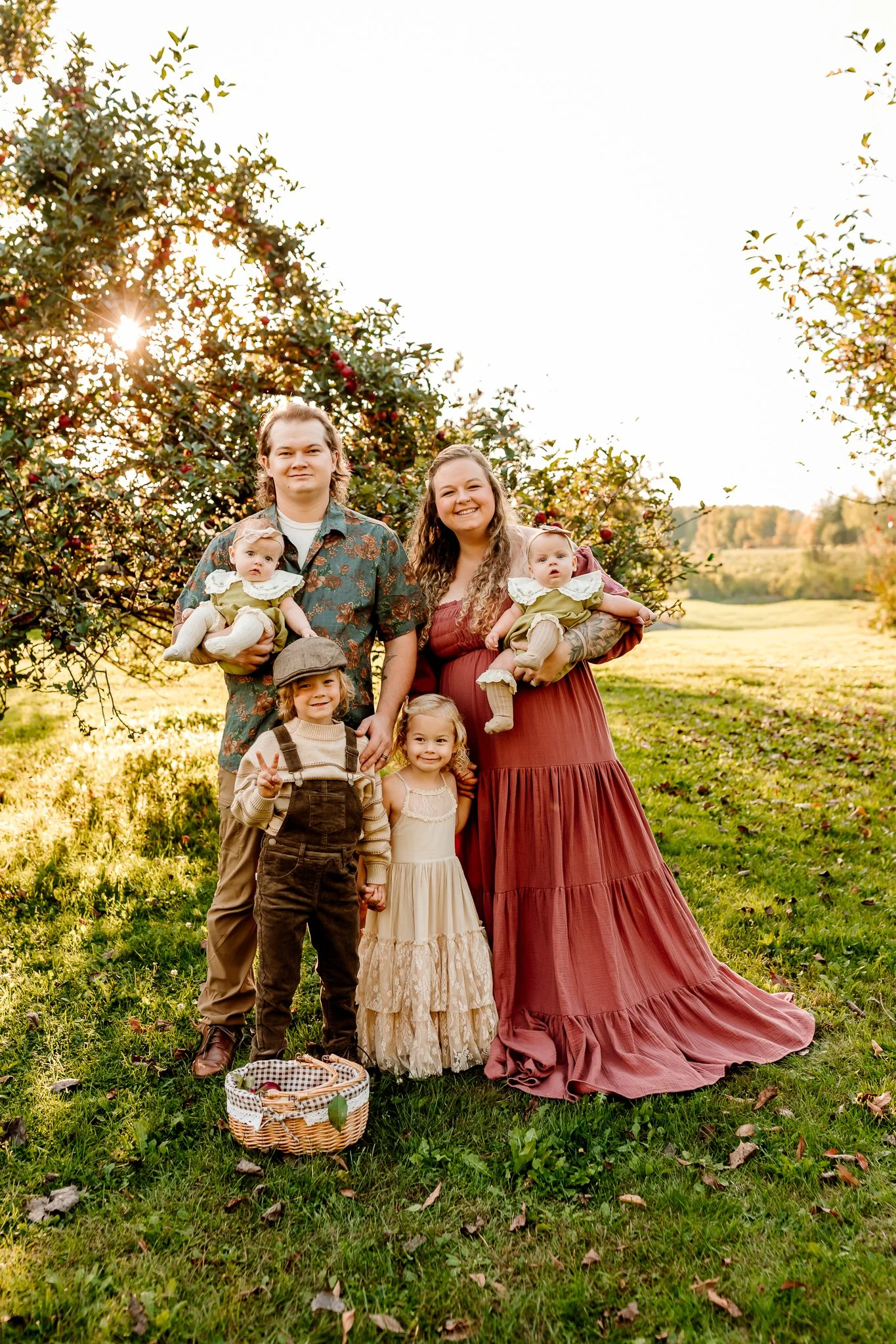 Family of six outdoors in a grassy field with trees, sun shining through leaves, and rolling hills in the background. Two women and four children, some holding babies, all smiling and dressed in vintage-style clothing.