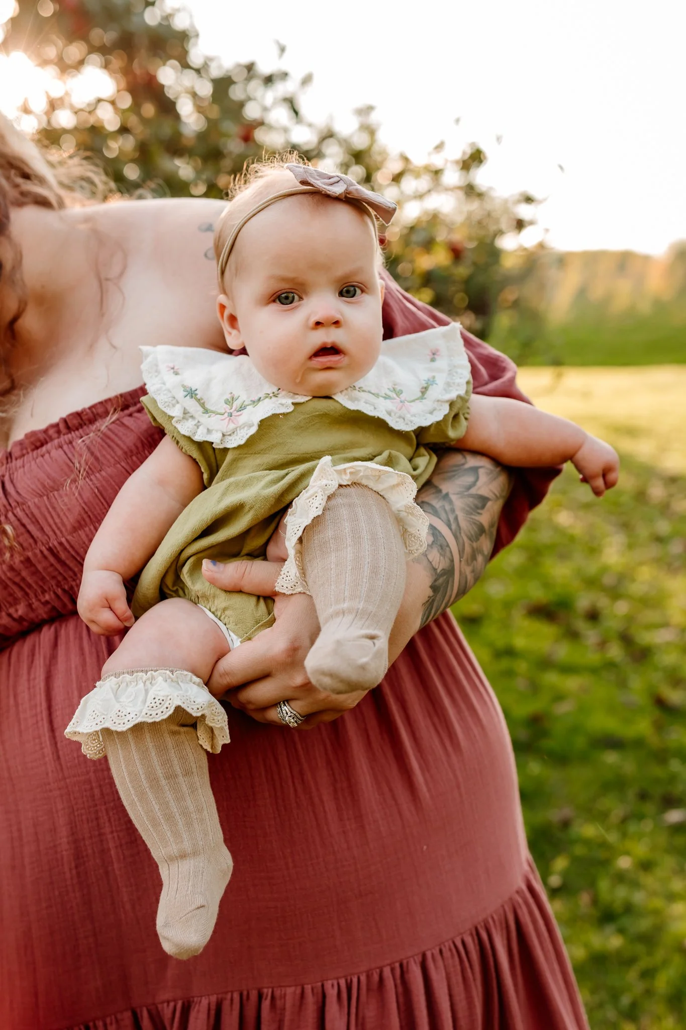 A baby girl with a surprised expression, wearing a green dress with embroidered flowers, cream-colored lace socks, and a beige headband with a bow, held by a person with tattoos on their arm outdoors during sunset with trees in the background.