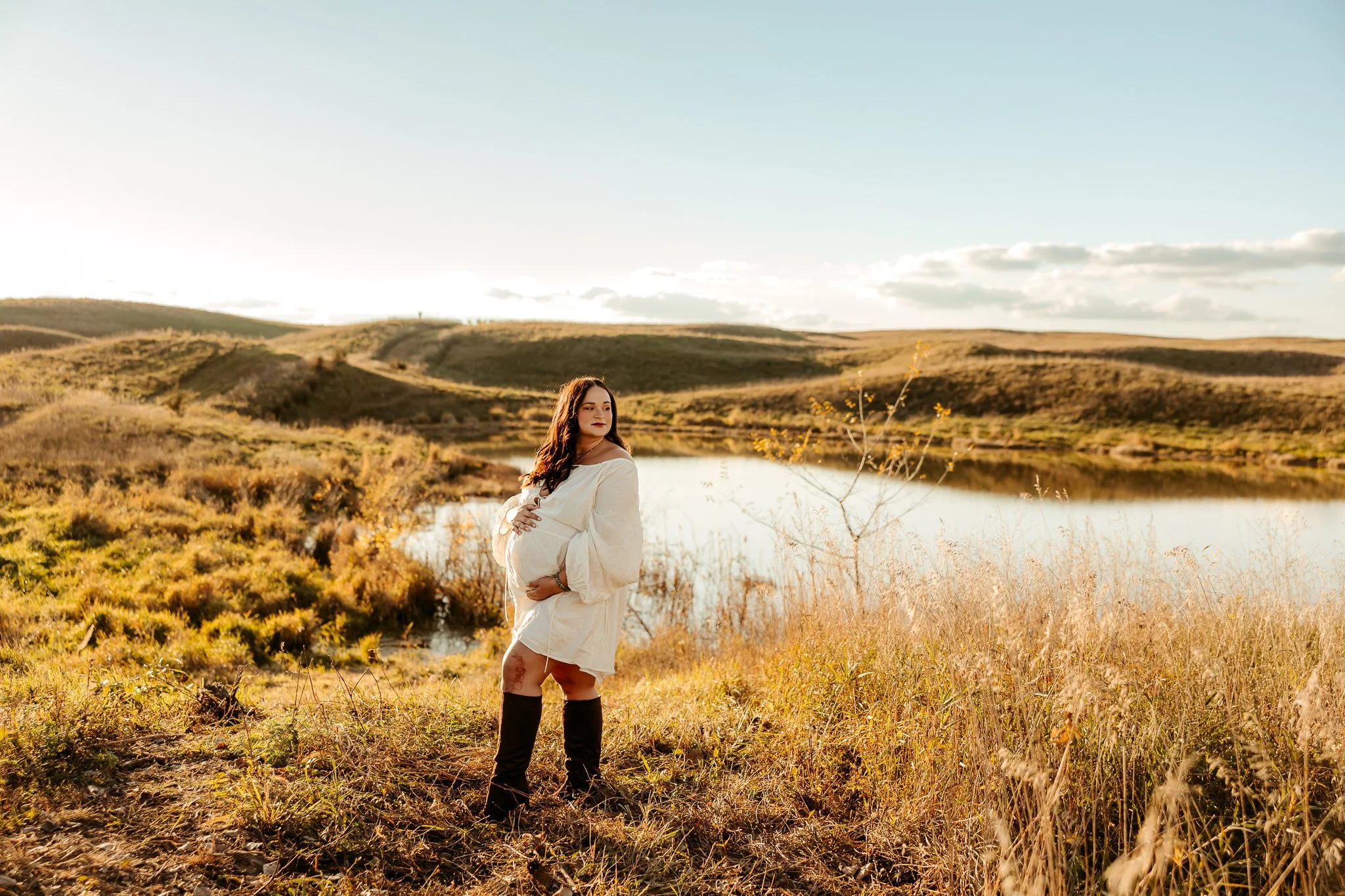 Pregnant woman standing outdoors in a grassy field near a body of water, with rolling hills and a cloudy sky in the background.