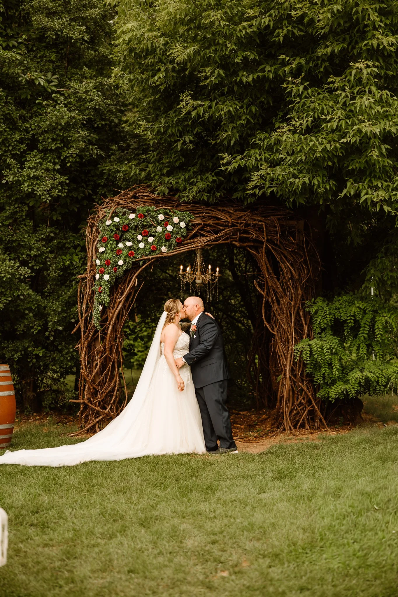 A bride and groom sharing a kiss during their outdoor wedding ceremony, standing under a rustic wooden arch decorated with flowers and greenery, with trees in the background.