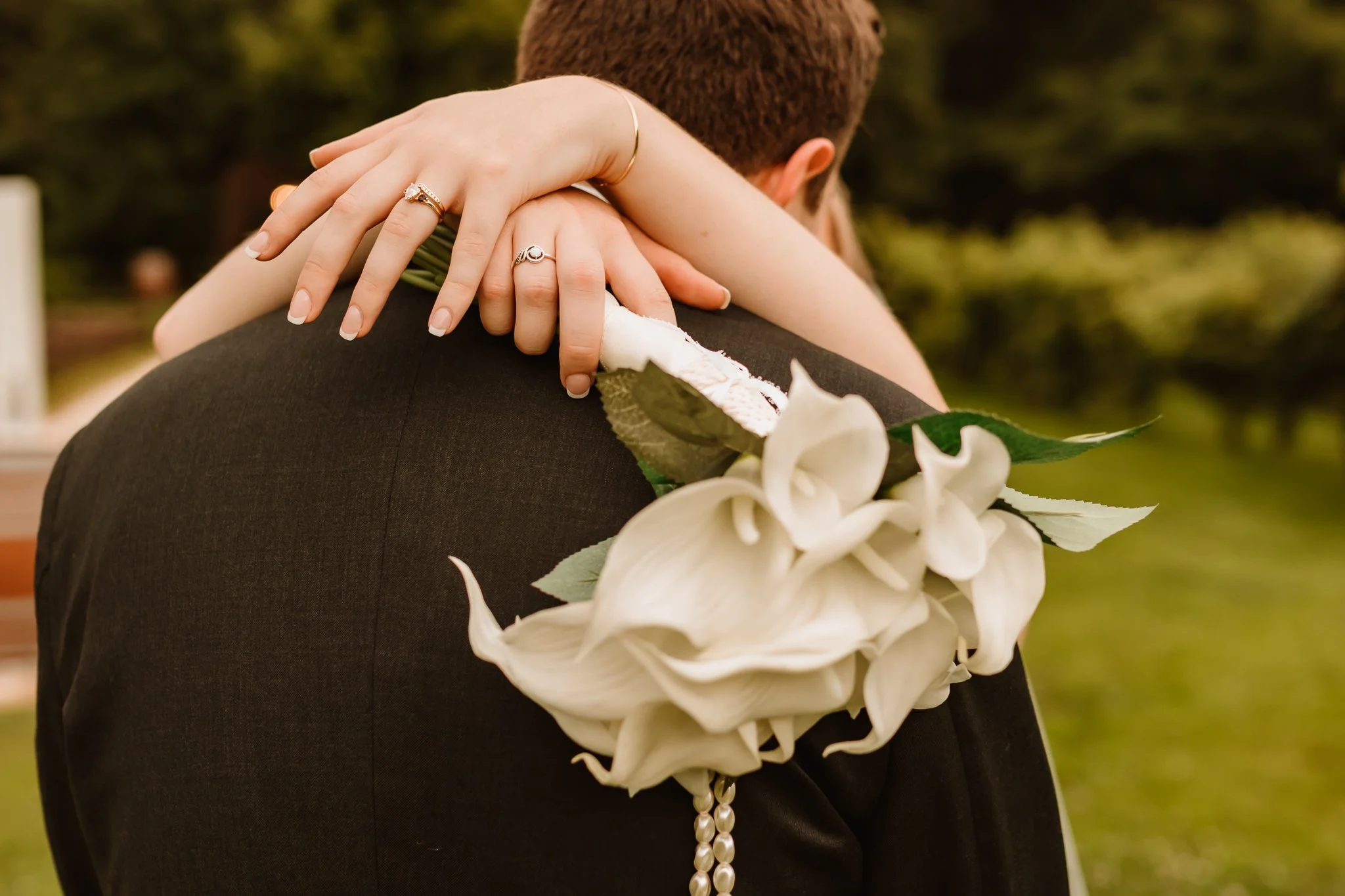 A bride and groom embrace, with the bride's arms wrapped around the groom's shoulders, revealing wedding rings. The bride's hand rests on the groom's shoulder, showing rings and a bracelet. The groom wears a dark suit and has a white floral boutonnie