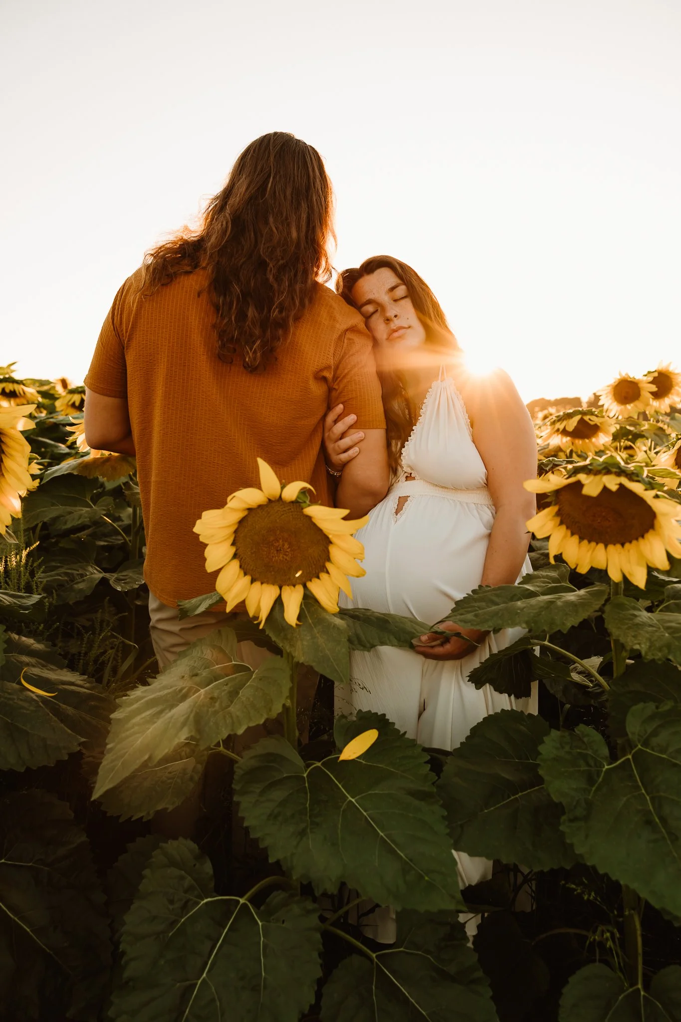 A pregnant woman in a white dress resting her head on a man's shoulder while standing among sunflowers at sunset.