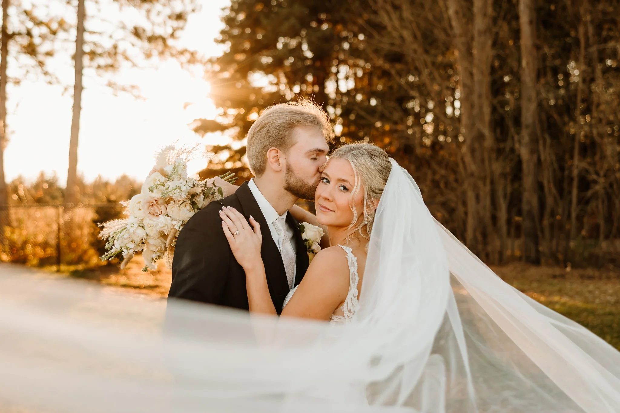 A newlywed couple is outdoors during sunset, with the groom kissing the bride on her temple. The bride is smiling softly, wearing a white wedding dress and veil, and holding a bouquet of flowers. The groom is wearing a black suit and white shirt. The