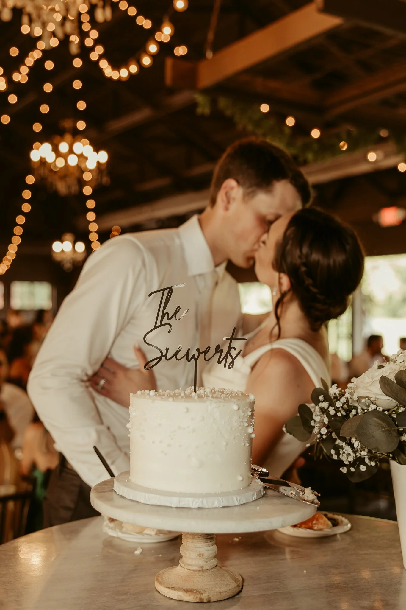 A newlywed couple sharing a kiss at their wedding reception, with a white wedding cake topped with a black 'The Suverts' cake topper in the foreground.