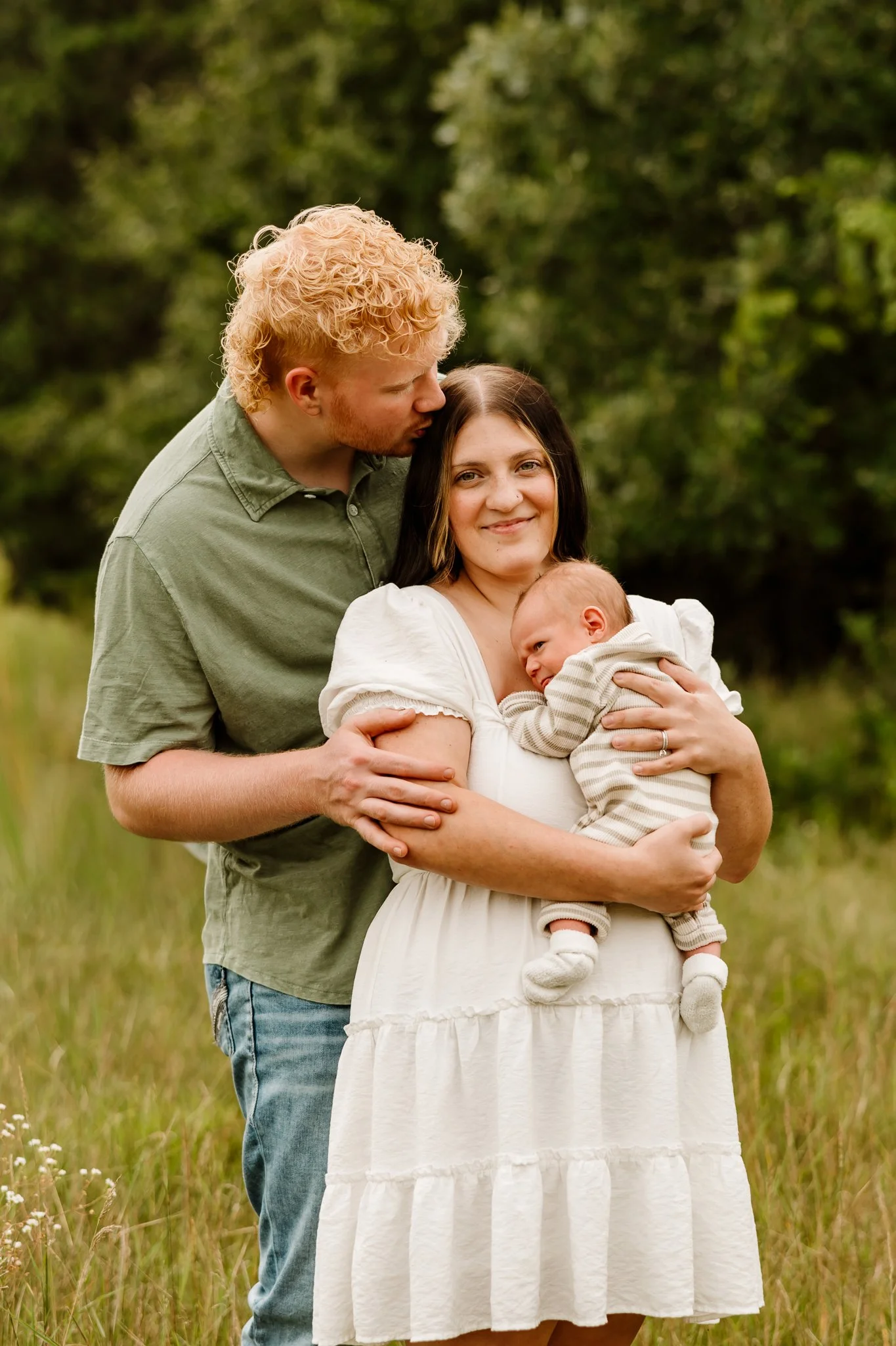 A family of three standing in a grassy field with trees in the background. The woman is holding a baby, and a man is kissing her on the forehead.