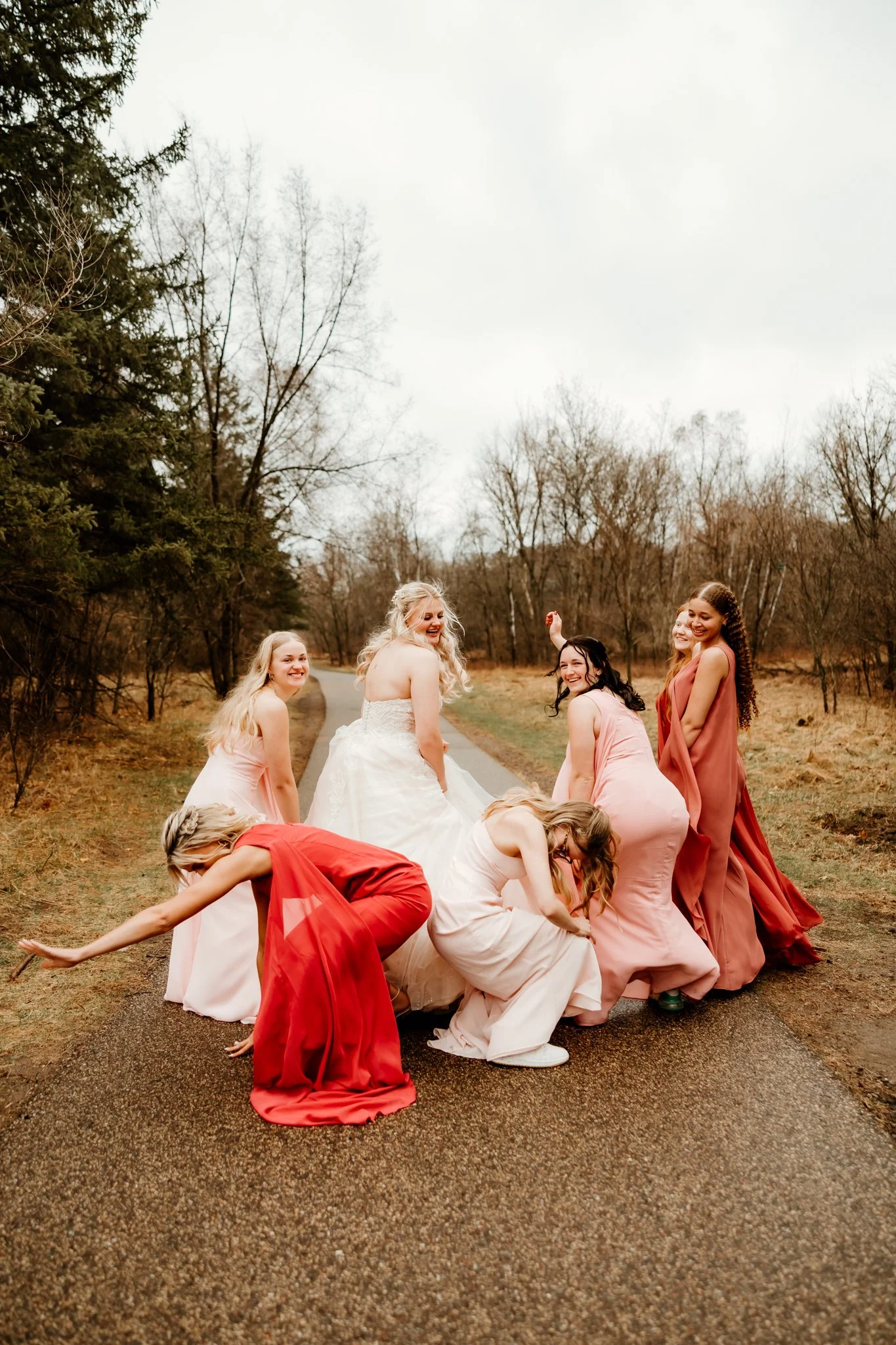 Group of women, including a bride in a wedding dress, posing playfully on a forested road in an outdoor setting.