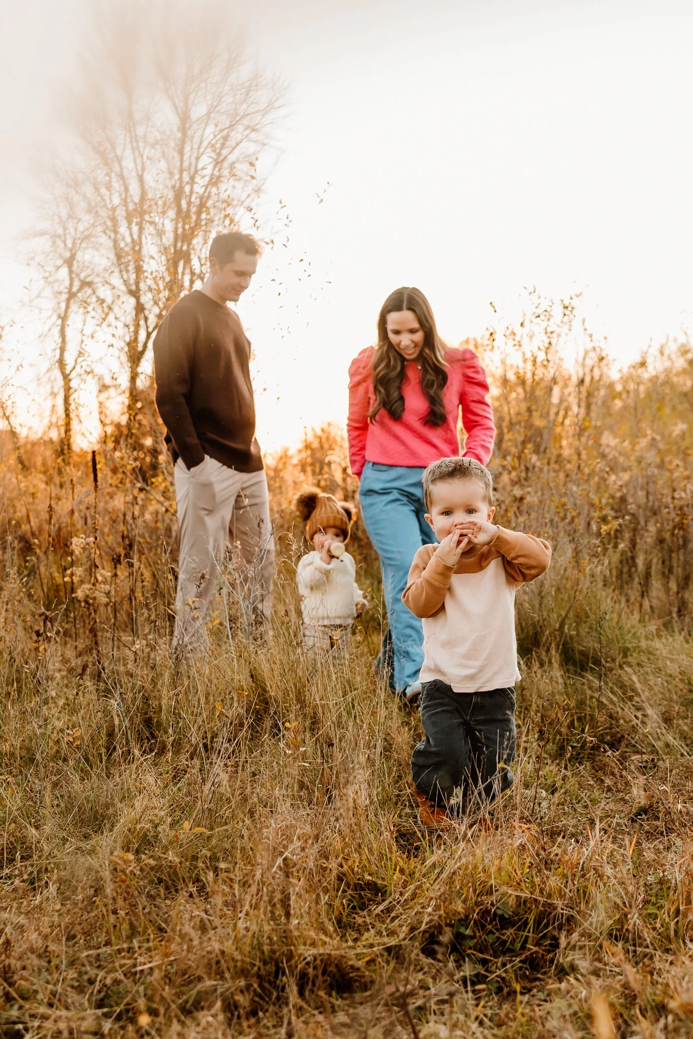 A family of four walking through a grassy field during autumn sunset, with bare trees in the background. The young boy in the foreground is wearing a beige and brown shirt, while the woman and man behind him are dressed in colorful clothing, and a yo