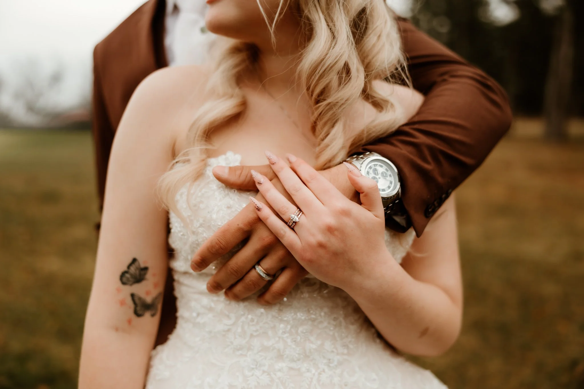 A woman in a wedding dress and a man in a brown suit embrace outdoors, showing her diamond ring and a tattoo of butterflies on her arm.