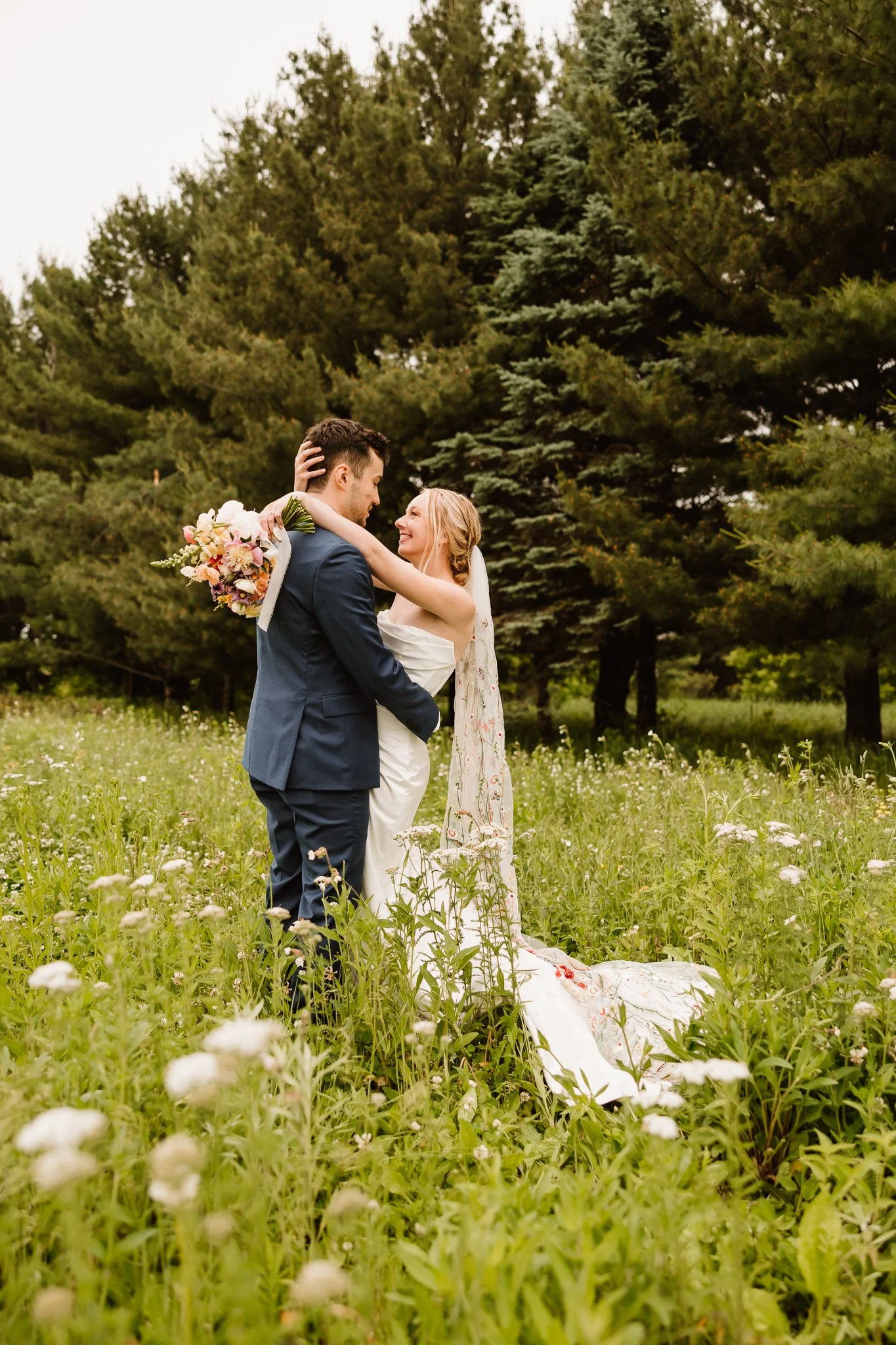 A bride and groom in a green field with trees, sharing a romantic moment, with the bride holding a bouquet of flowers.