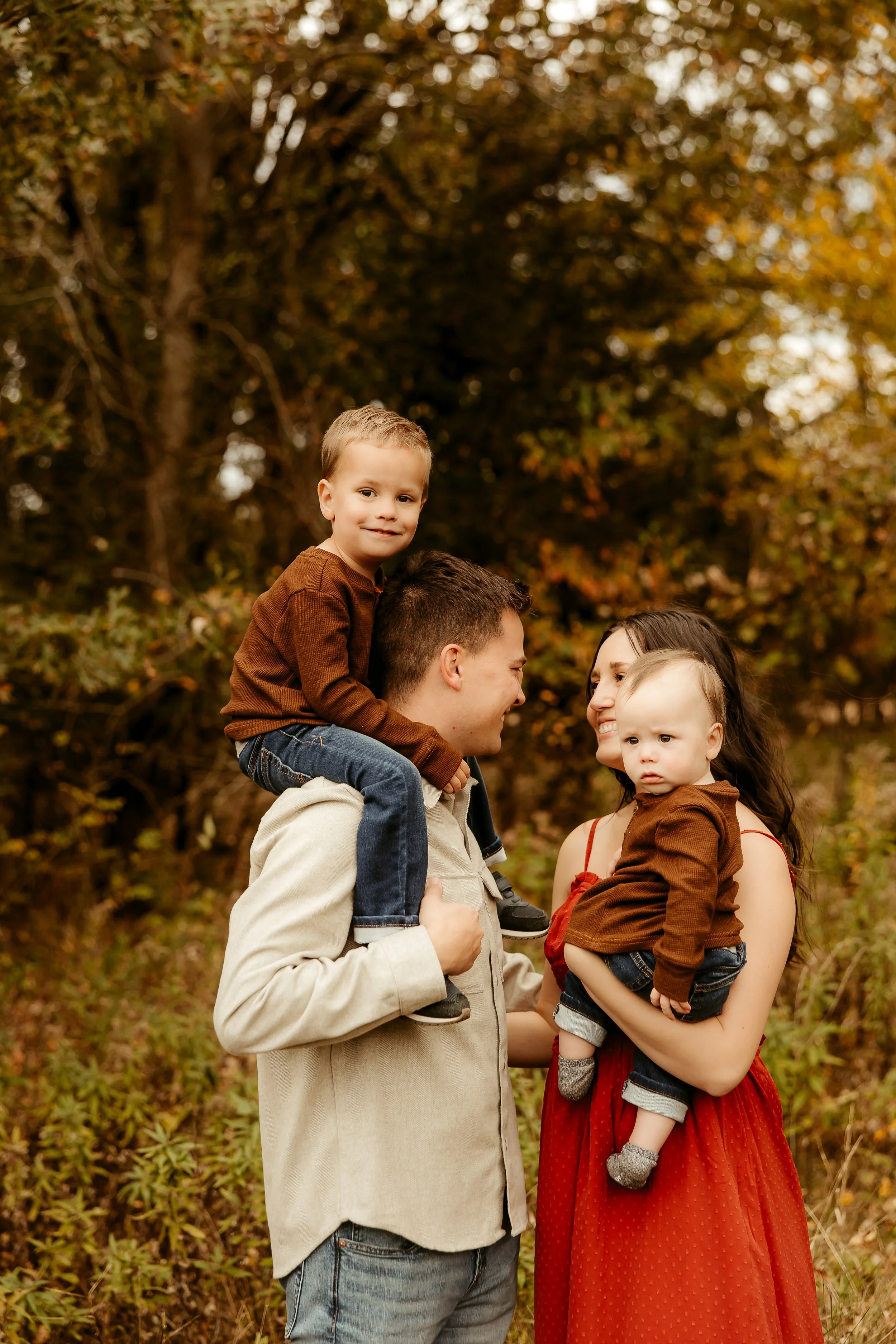 A family of four standing outdoors during autumn, with trees and fall foliage in the background. The father, wearing a beige jacket, carries a young boy on his shoulders, while the mother, in a red dress, holds a toddler.Minnesota family photographer