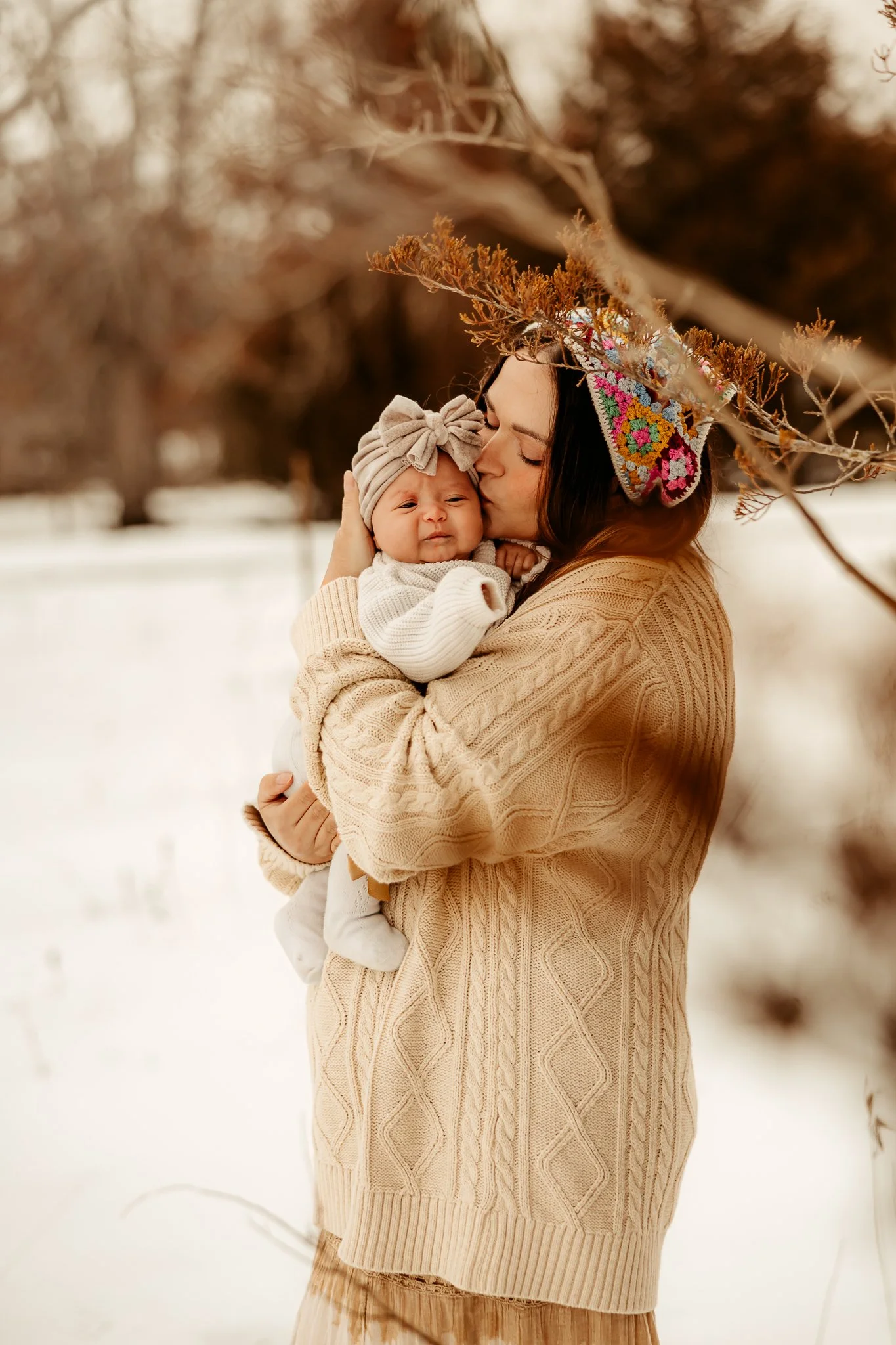 A woman holding a baby in a snowy outdoor setting, woman wearing a beige sweater and kissin baby's forehead, under bare trees.