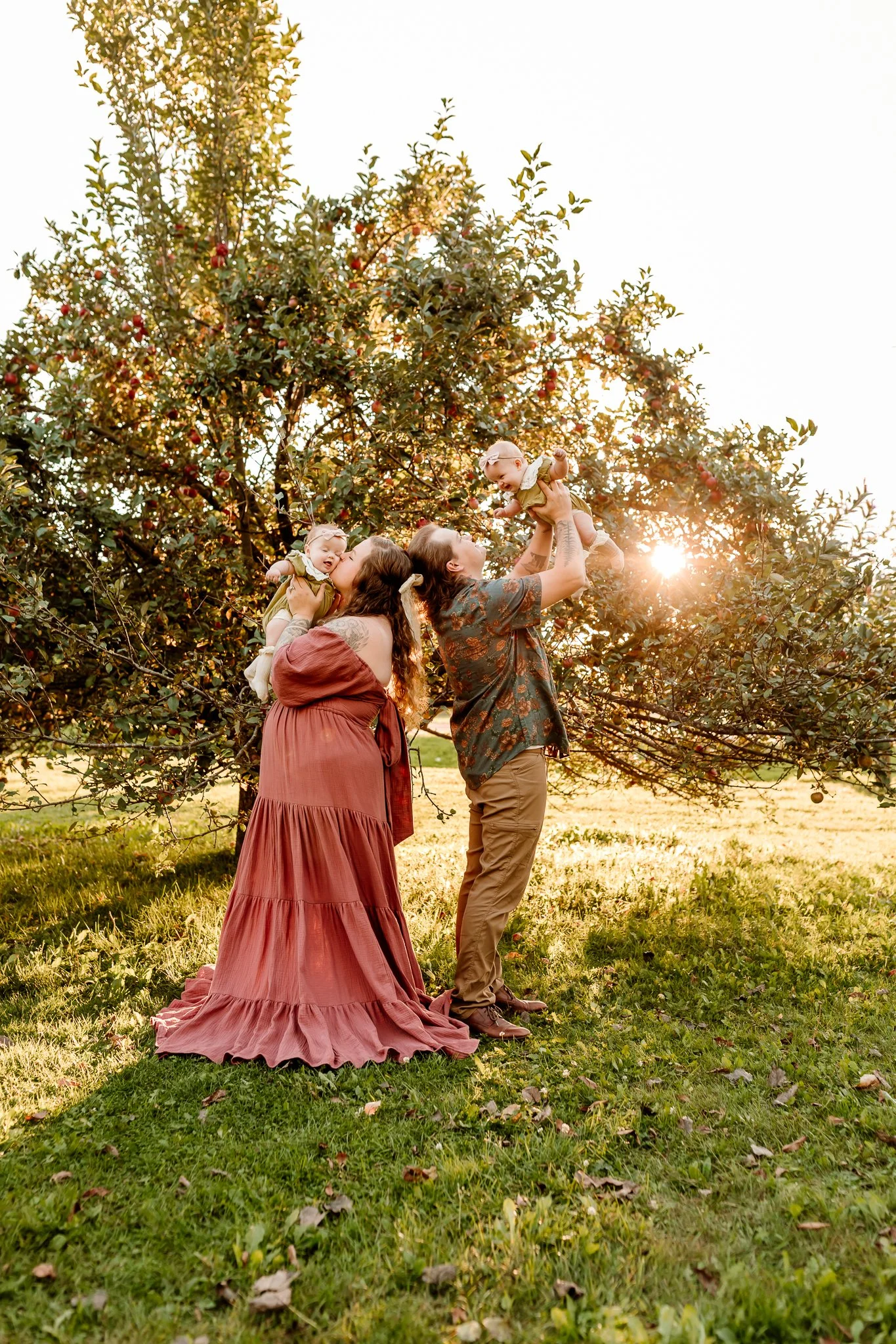 A family of four enjoying a sunny day outdoors, with the father lifting a baby girl in front of a large apple tree, while the mother holds another baby girl, all standing on grass.