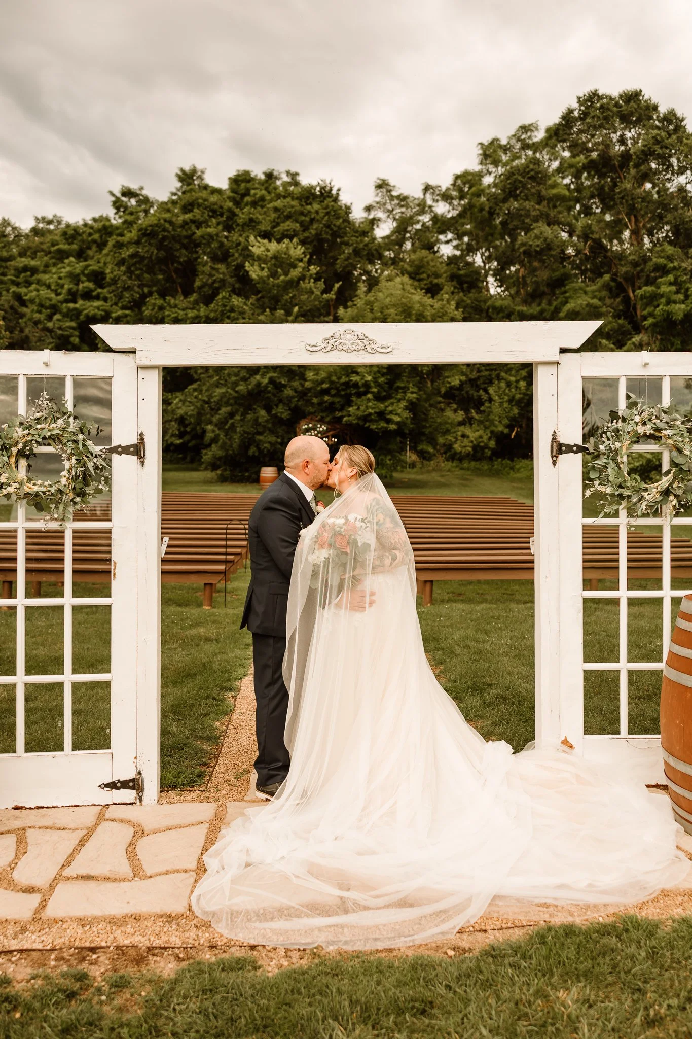 A bride and groom sharing a kiss underneath a white wooden wedding arch outdoors, with greenery and a cloudy sky in the background. Hope Glen Farm Cottage Grove Minnesota 