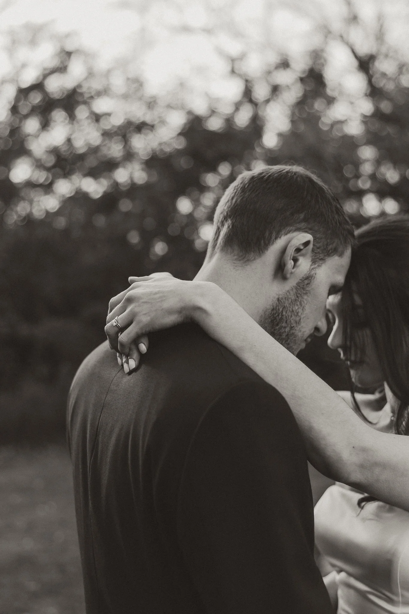 A close-up black and white photo of a couple embracing with their foreheads touching outdoors, blurred trees in the background.