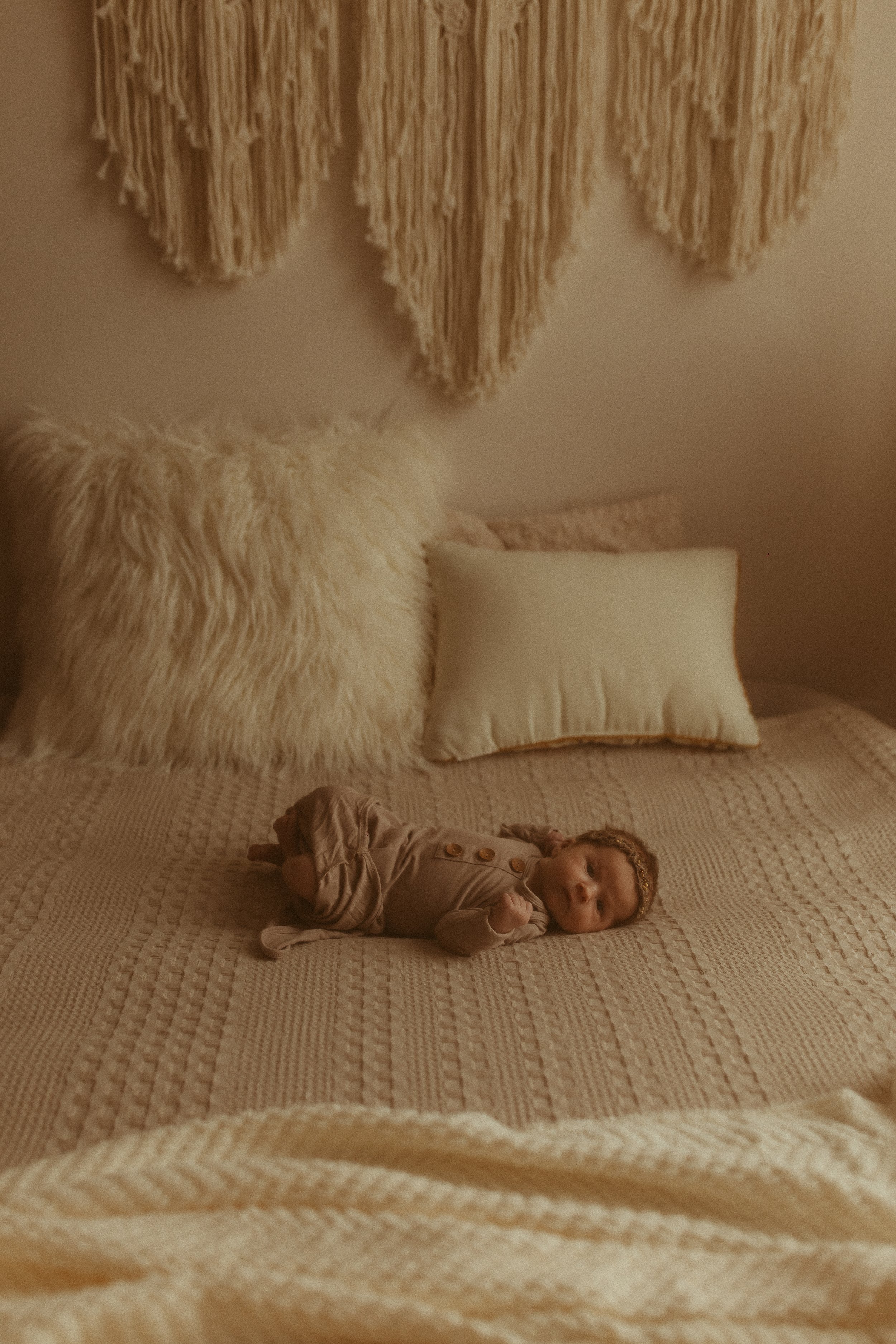 A baby lying on a textured beige bedspread surrounded by cozy pillows, with a large, cream-colored fur pillow and a small rectangular pillow nearby, above a decorative wall hanging.