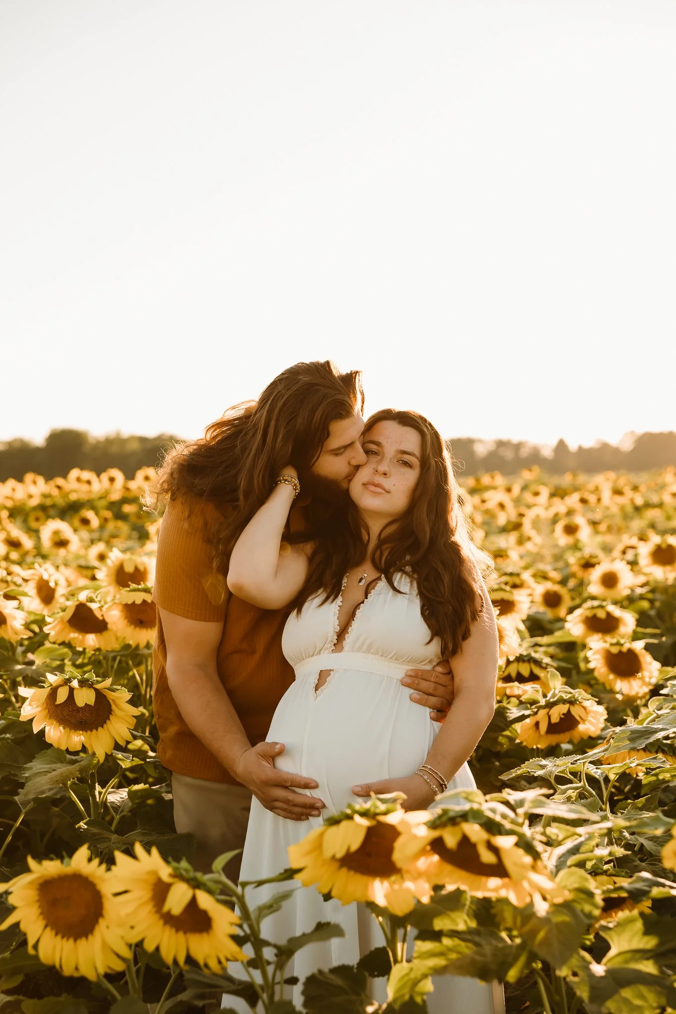 A pregnant woman in a white dress standing in a sunflower field, with a man embracing her from behind and kissing her on the cheek during sunset.