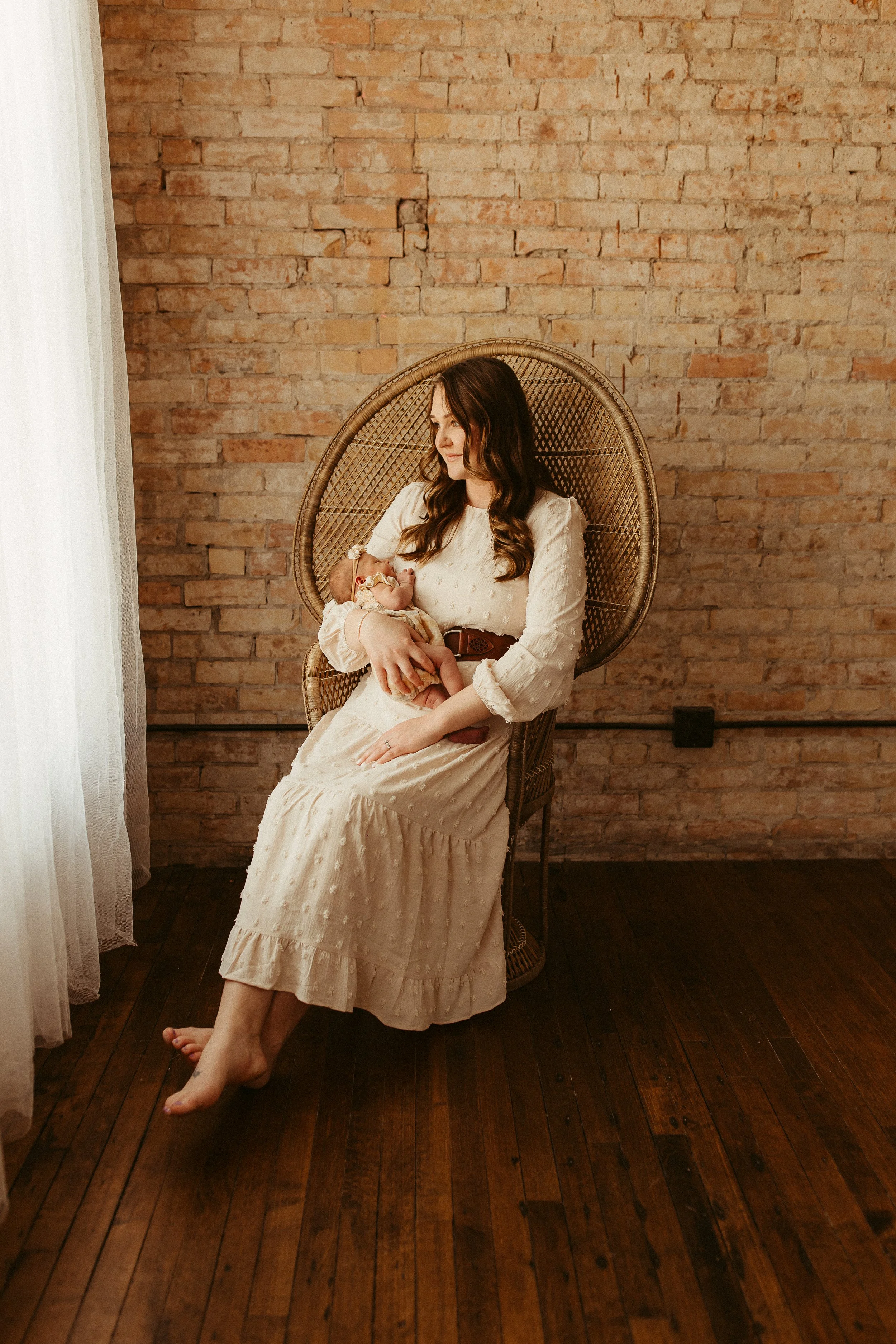 A woman sitting on a wicker chair holding a newborn baby in her arms, in a room with a brick wall and wooden floor.